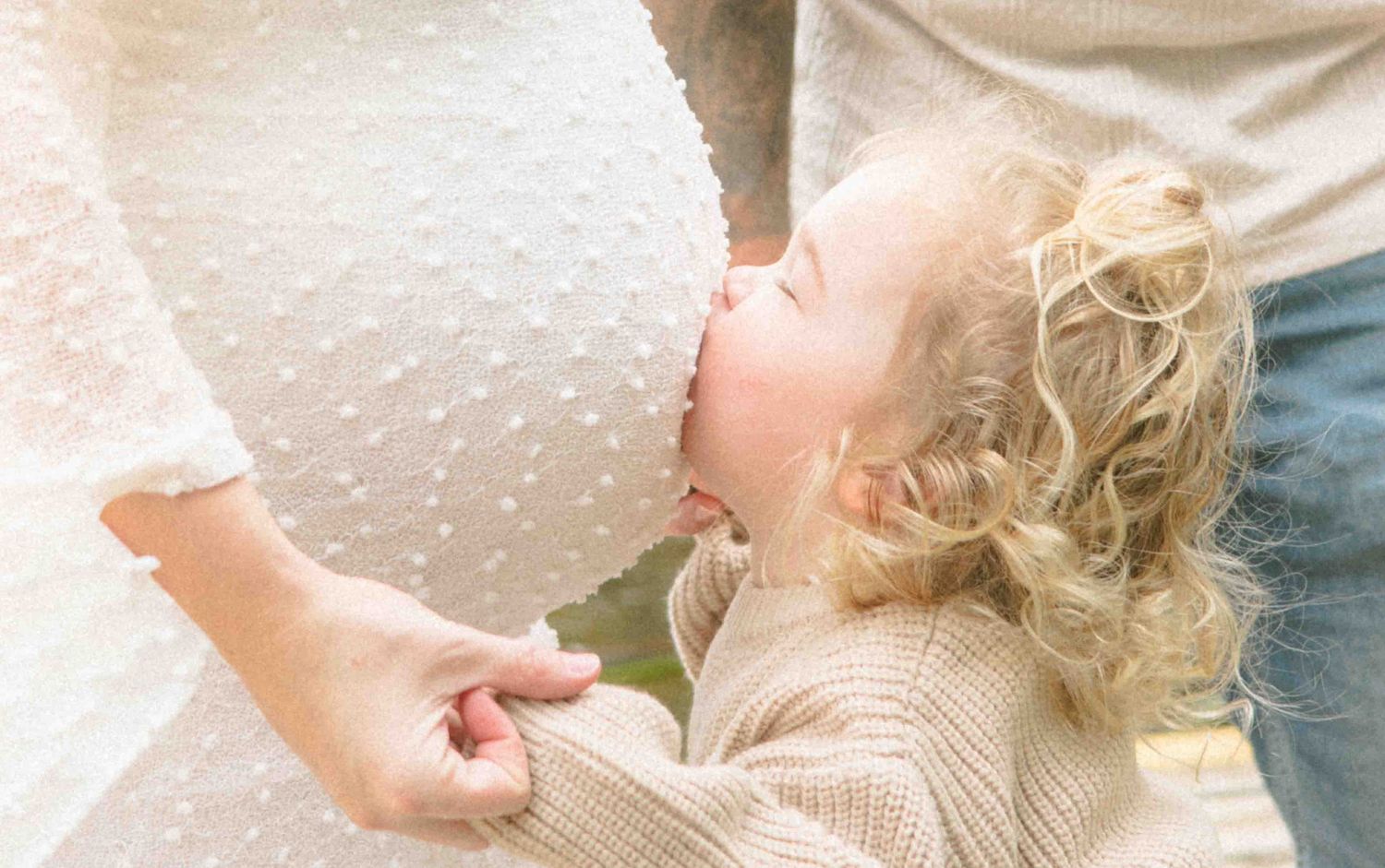 Toddler with curly blonde hair kisses the pregnant belly of a mother in a white dotted dress during an outdoor maternity photo session.