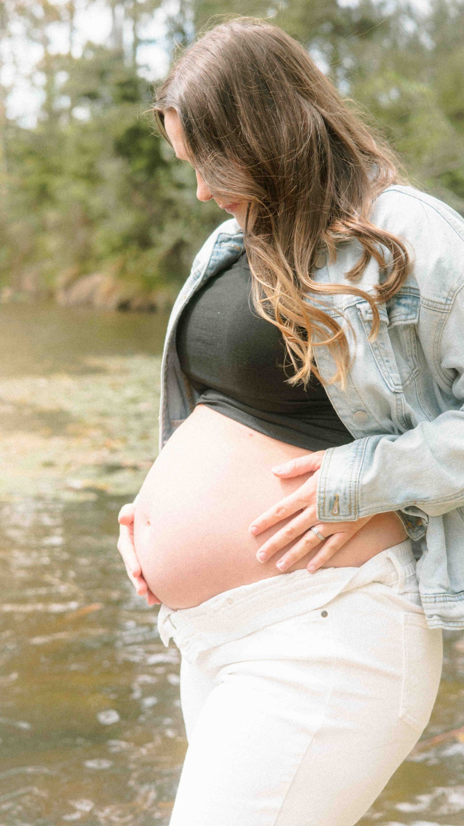 Pregnant woman in a denim jacket and white pants cradles her bare baby bump while standing beside a calm river in a maternity photoshoot.