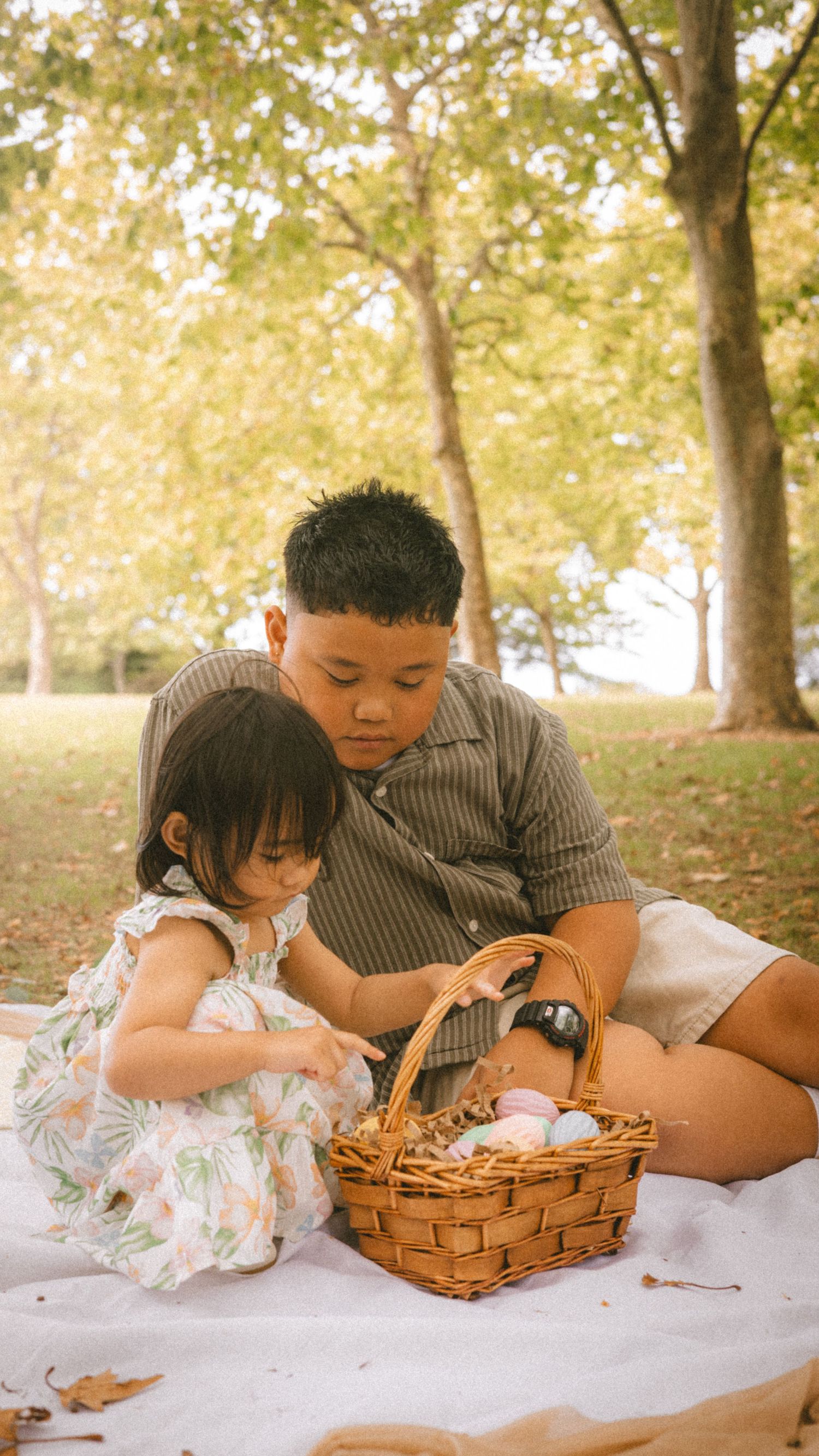 Two children sit on a blanket in a park, looking at a wicker Easter basket filled with eggs.