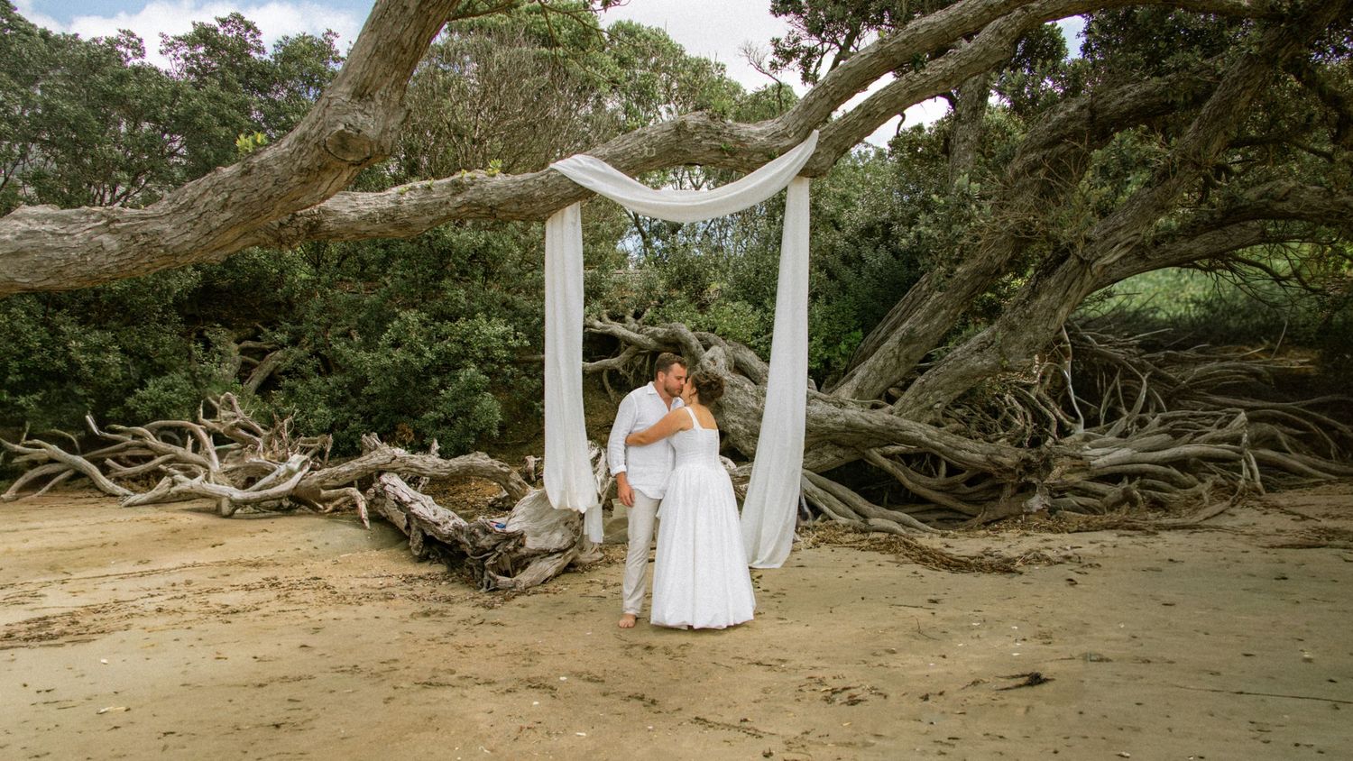 Couple embracing in wedding attire on sandy beach beneath rustic arch with draped fabric and fallen tree backdrop.