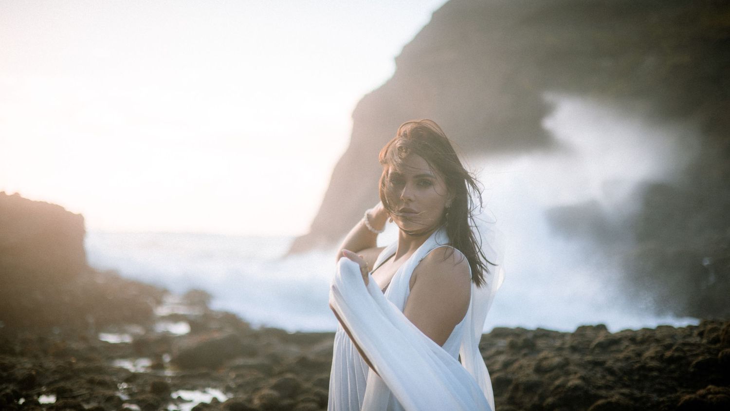 Artistic beach photoshoot featuring flowing white fabric against dramatic coastal scenery.