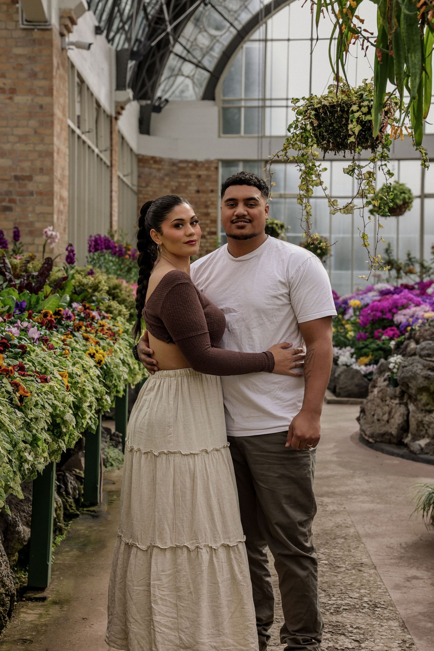 A romantic couple poses together in the winter gardens walkway surrounded by flowering plants and greenery.