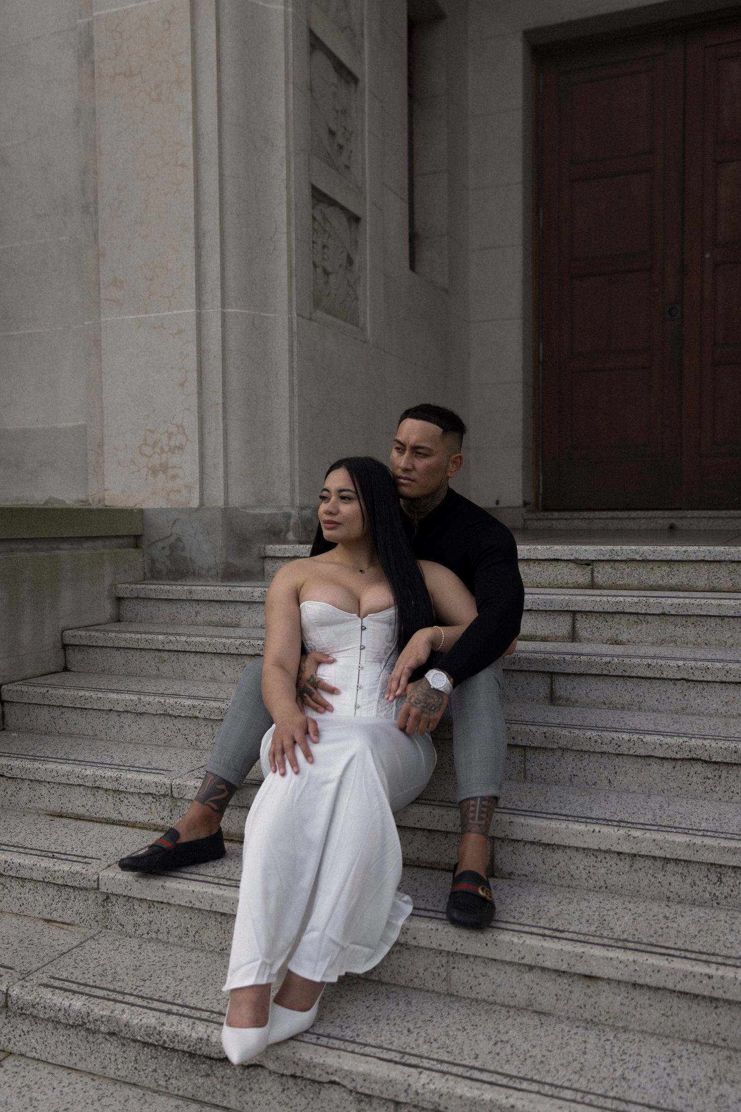 A couple in formal attire sits intimately on stone steps of a grand building during a romantic photoshoot.