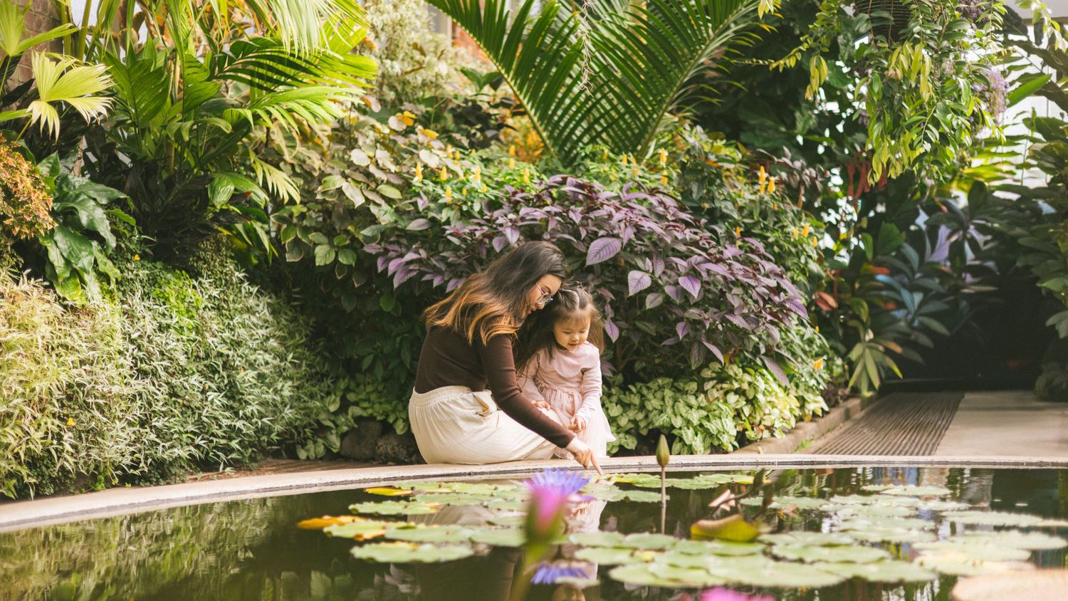A mother and child lean over a tranquil lily pond surrounded by lush tropical greenery in a botanical garden.