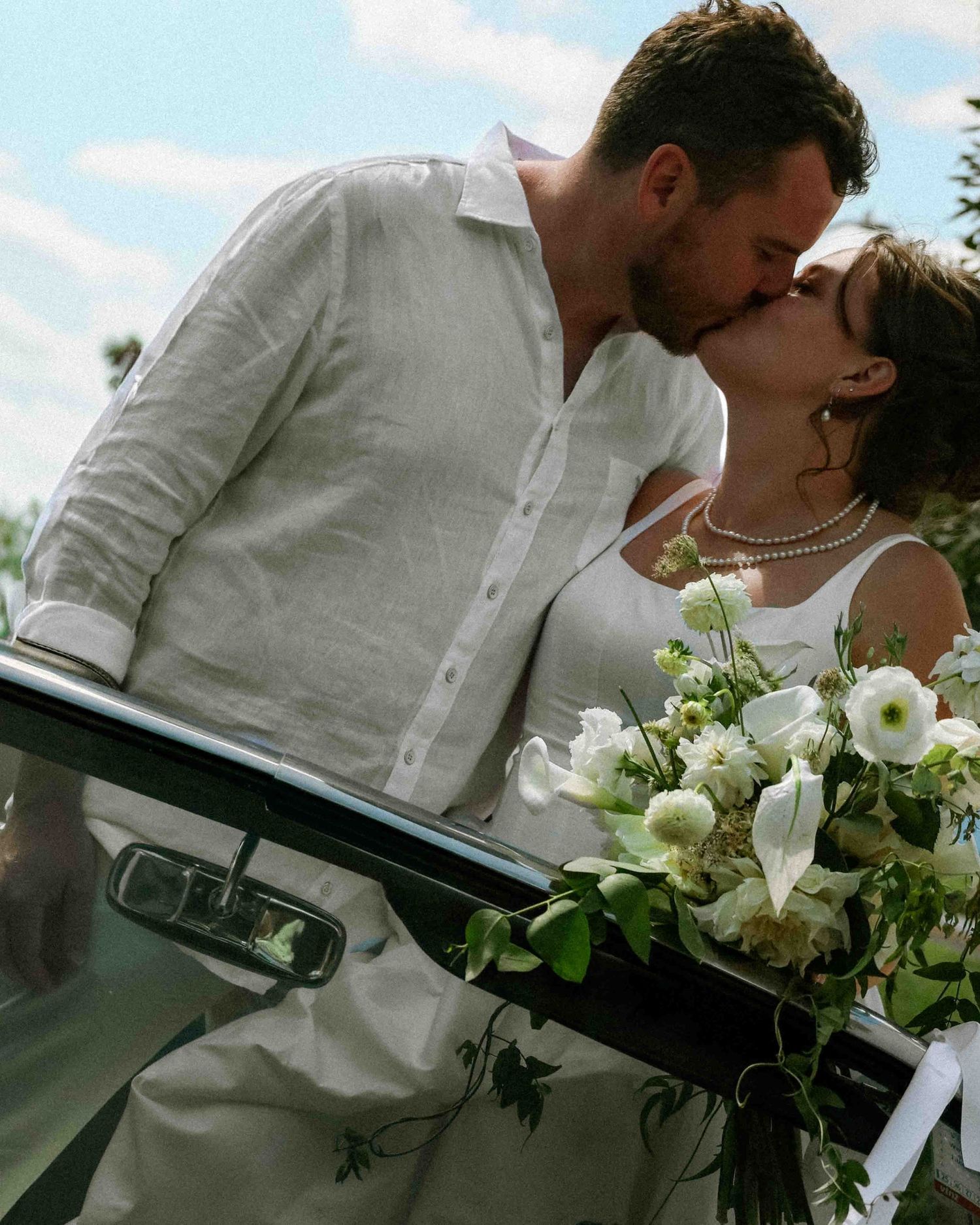 A bride and groom share a romantic kiss beside a vintage car, bride holding white floral bouquet on their wedding day.