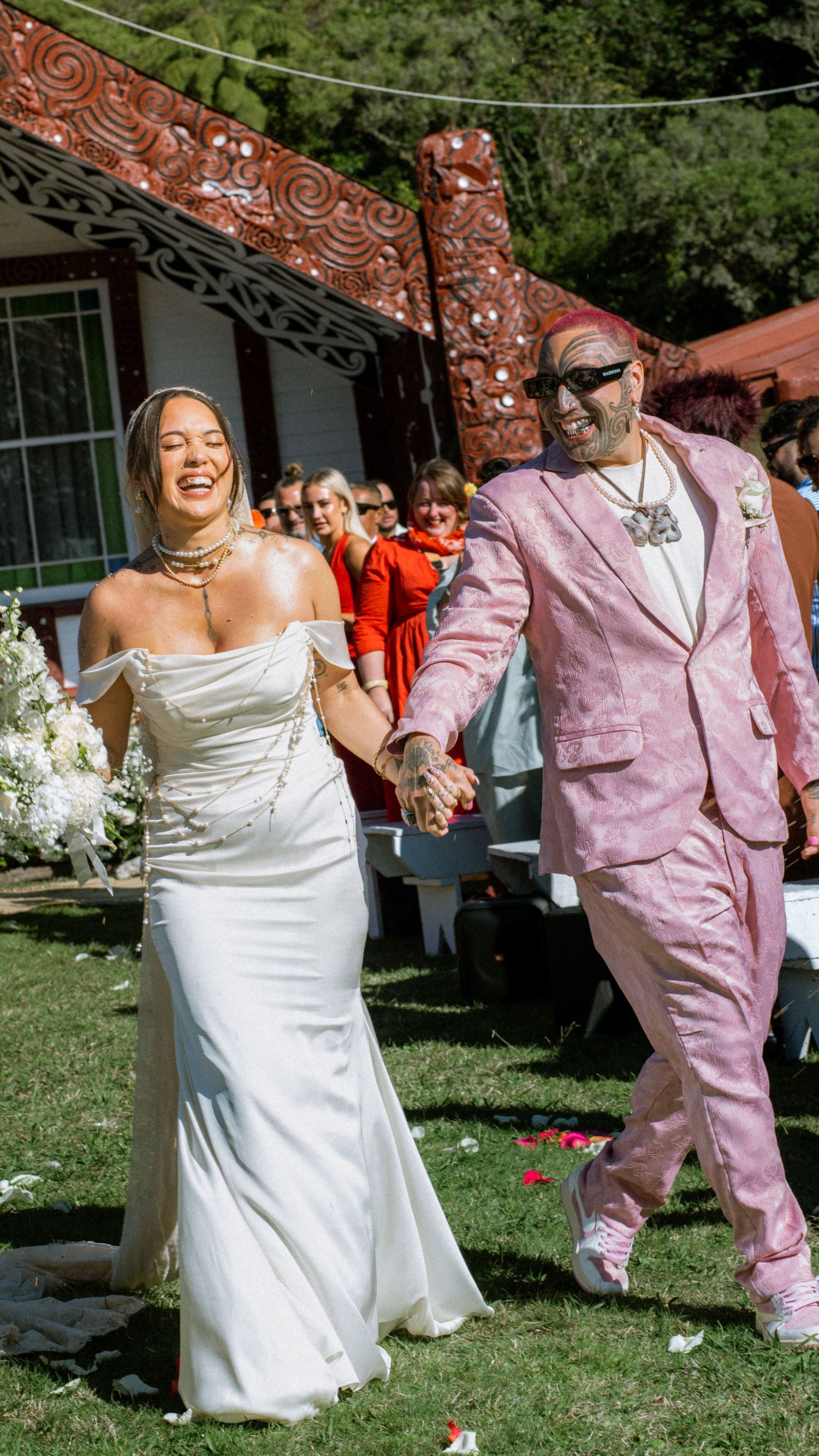 A wedding celebration on a lawn with a bride in white gown and partner wearing pink suit in front of traditional carved posts.