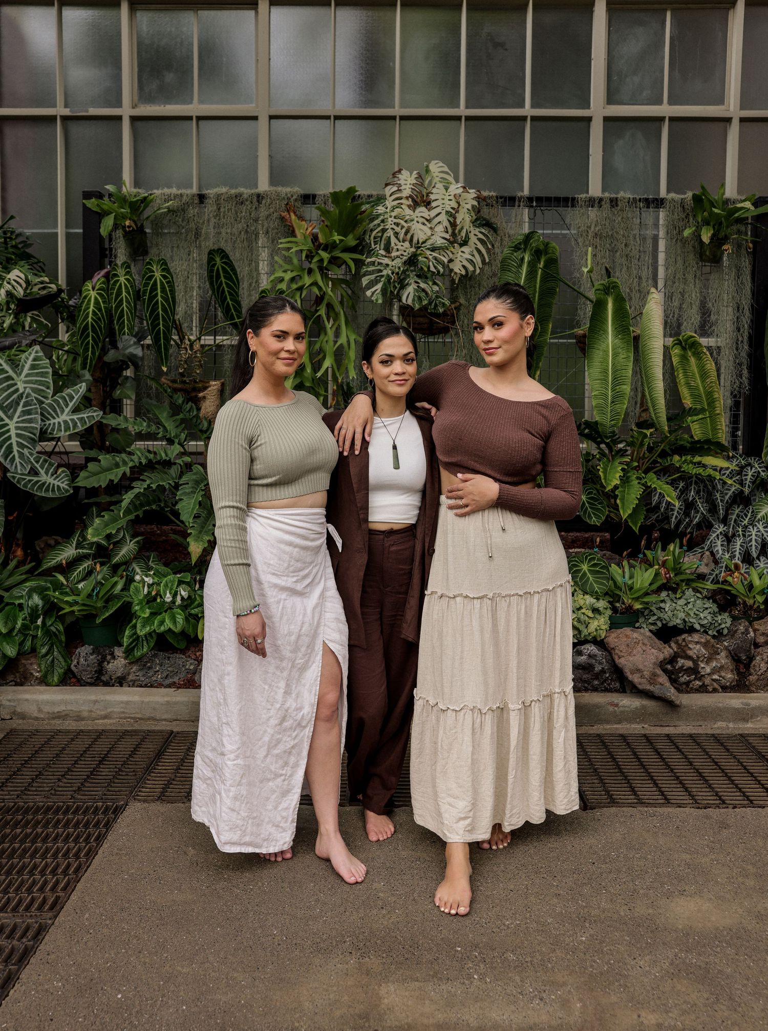 sisters in earth-toned dresses pose casually against a greenhouse wall.