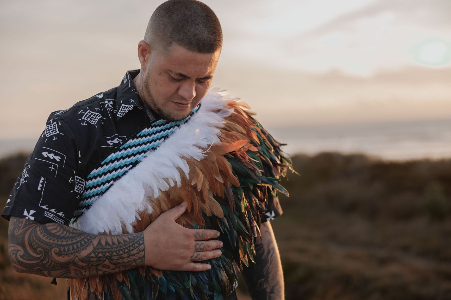 Person wearing traditional Polynesian clothing holds feathered ceremonial garment at sunset against natural landscape.