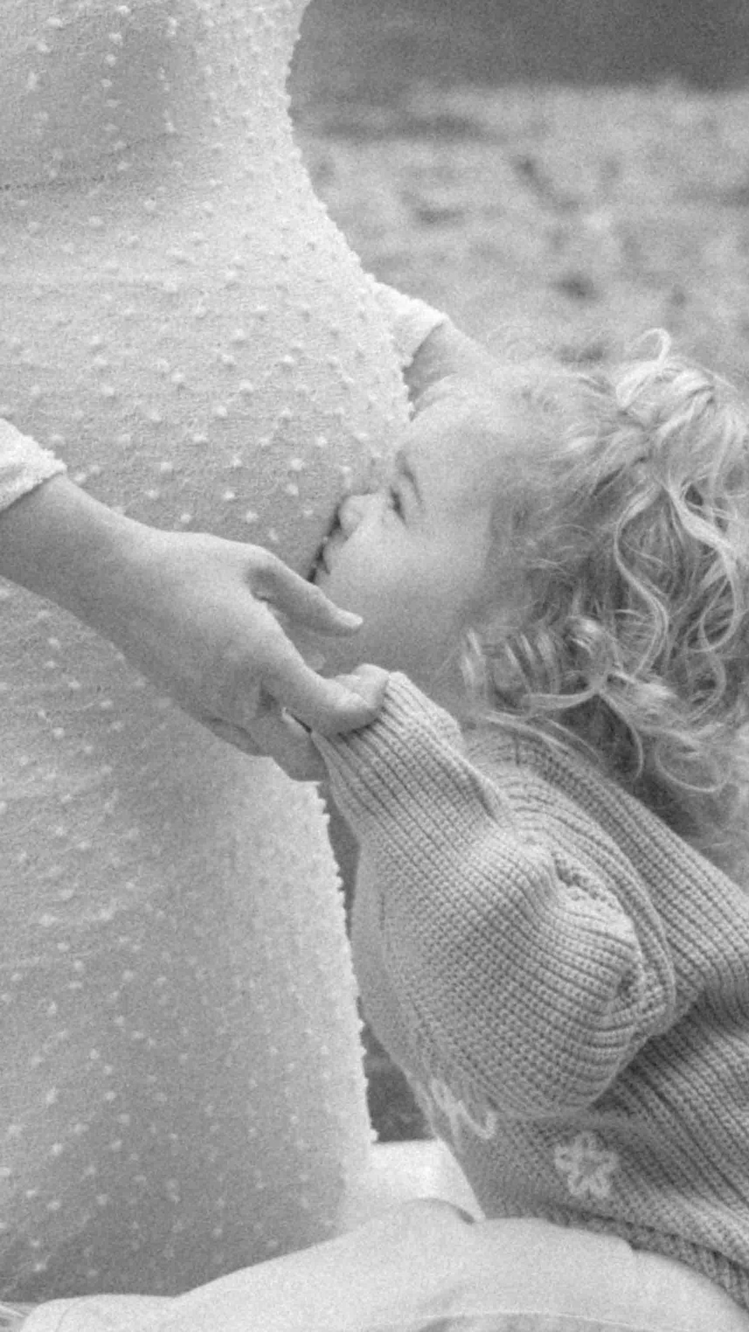Black and white photo of a curly-haired toddler in a knit cardigan tenderly kissing the belly of a pregnant woman in a dotted dress.