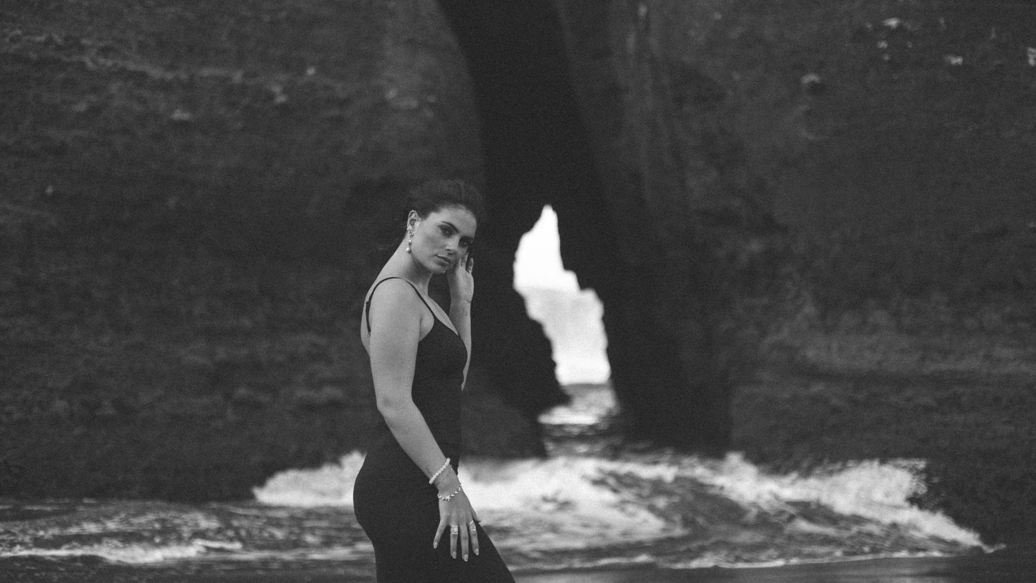 Artistic black and white photo of person posing near ocean waves crashing against rocks.
