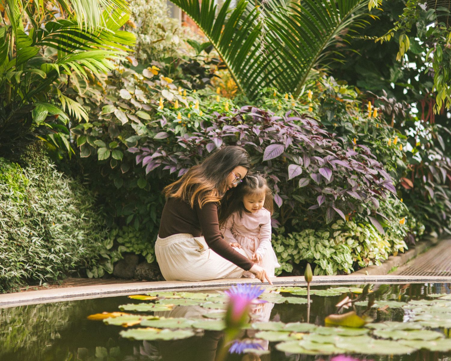 A mother and young daughter kneel by a serene lily pond surrounded by lush tropical greenery in a botanical garden.