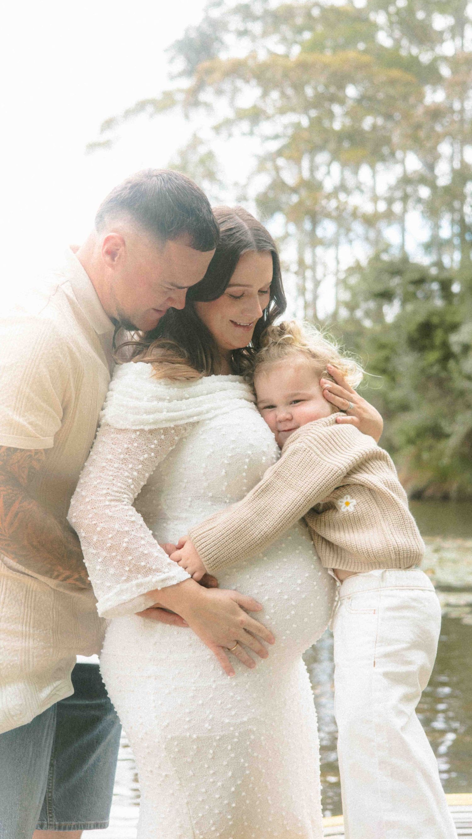 Expectant couple embracing in a lakeside maternity photo, the mother in a white dotted dress as their toddler hugs her baby bump.