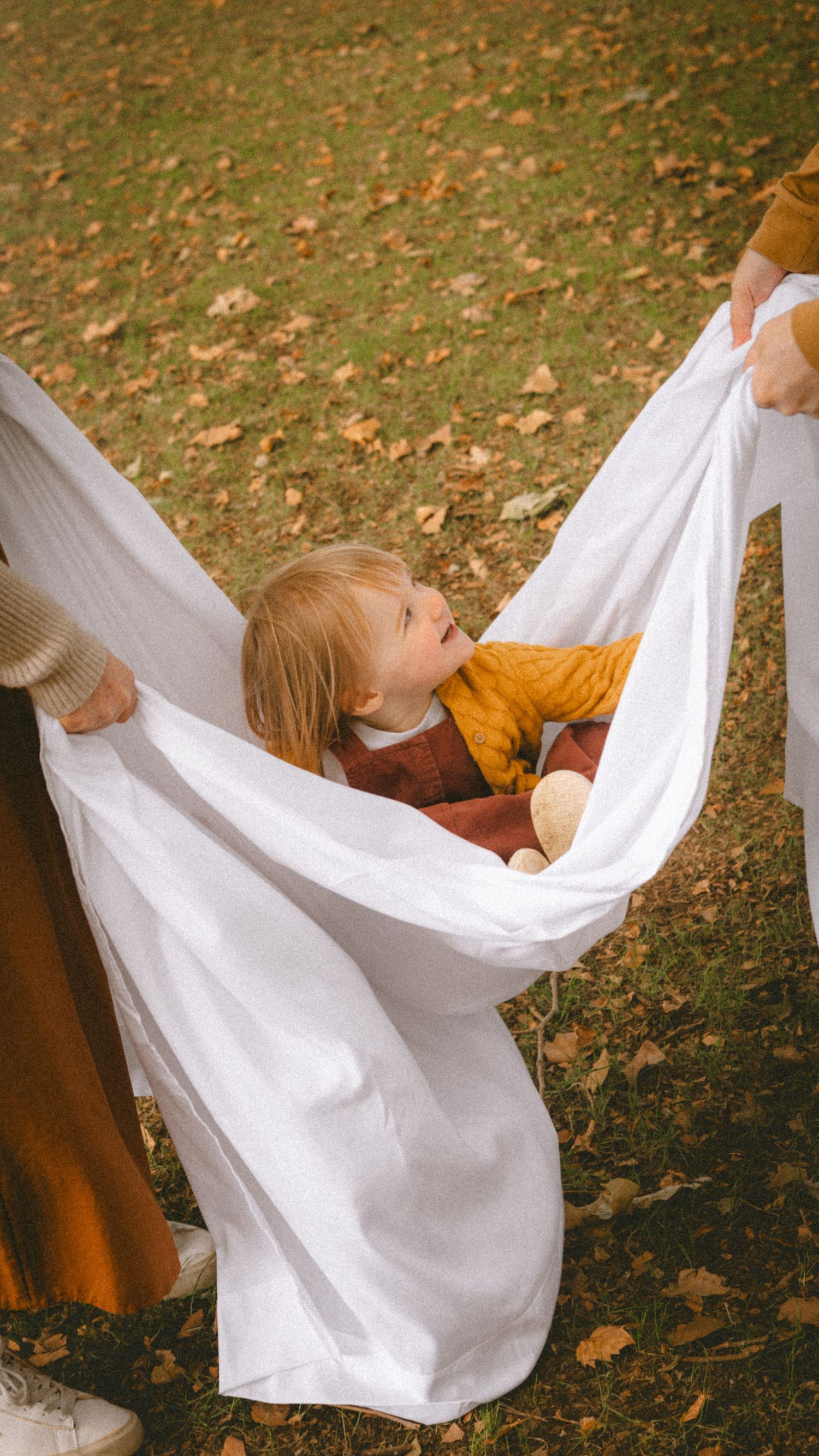 Toddler in mustard outfit cradled in white fabric hammock held by parents on autumn leaf-covered ground.