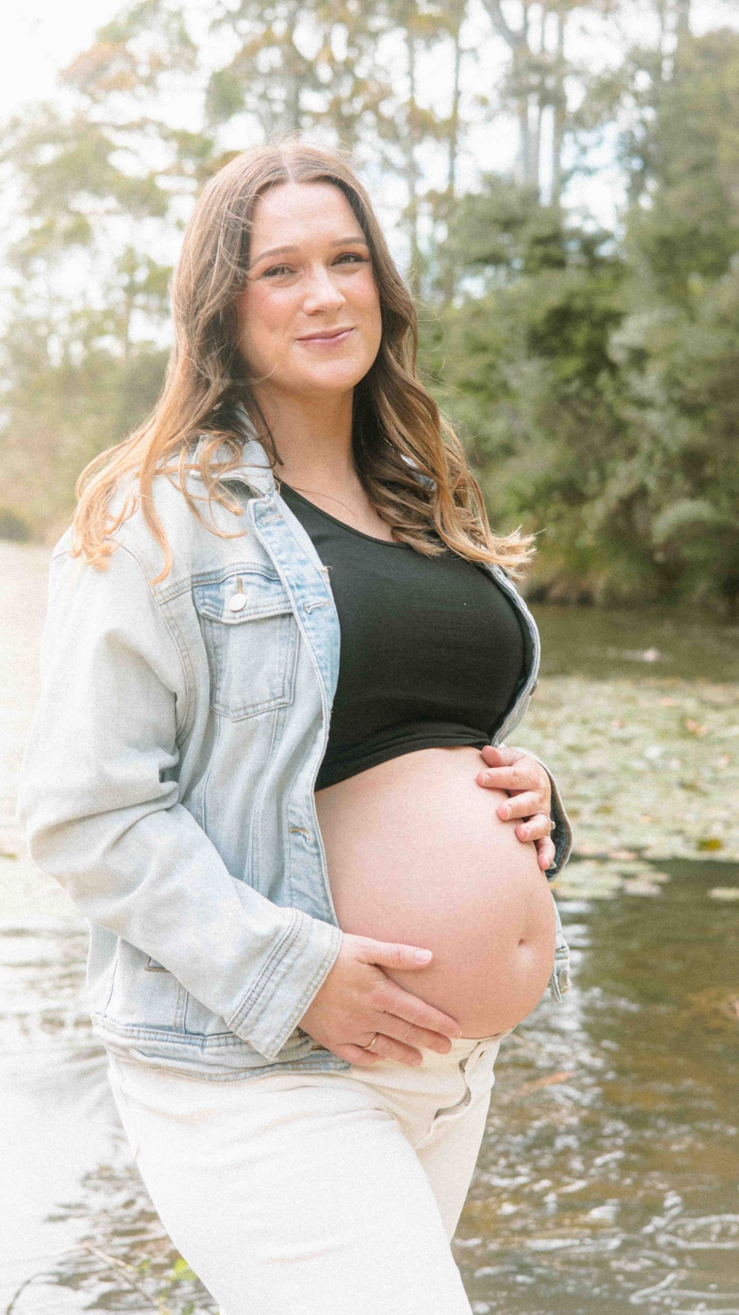 Pregnant woman in a denim jacket and black crop top cradles her bare baby bump beside a tranquil pond in a sunny, tree-lined park.