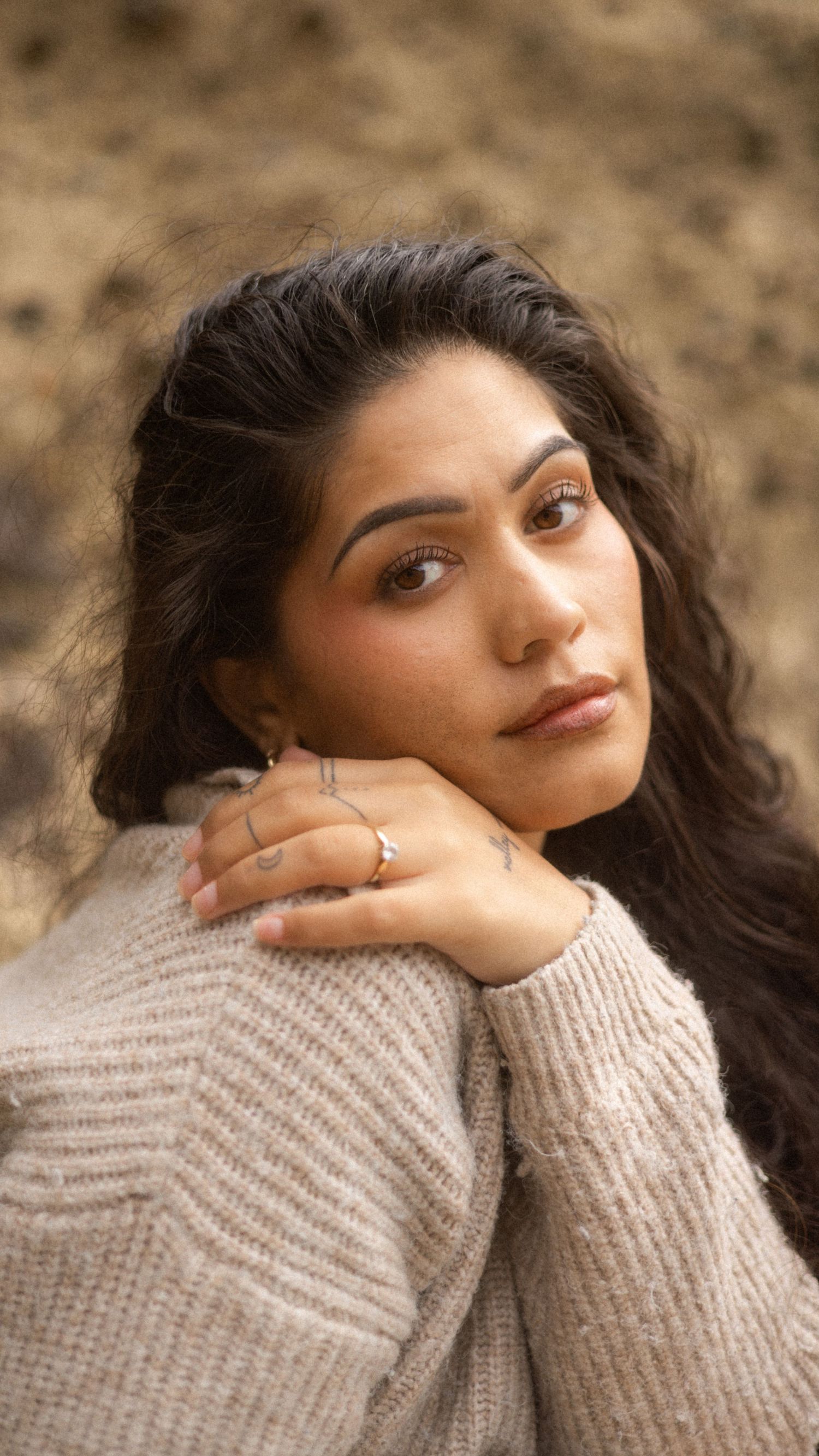 A person in a beige knit sweater poses thoughtfully with their hand on their face against a blurred background.