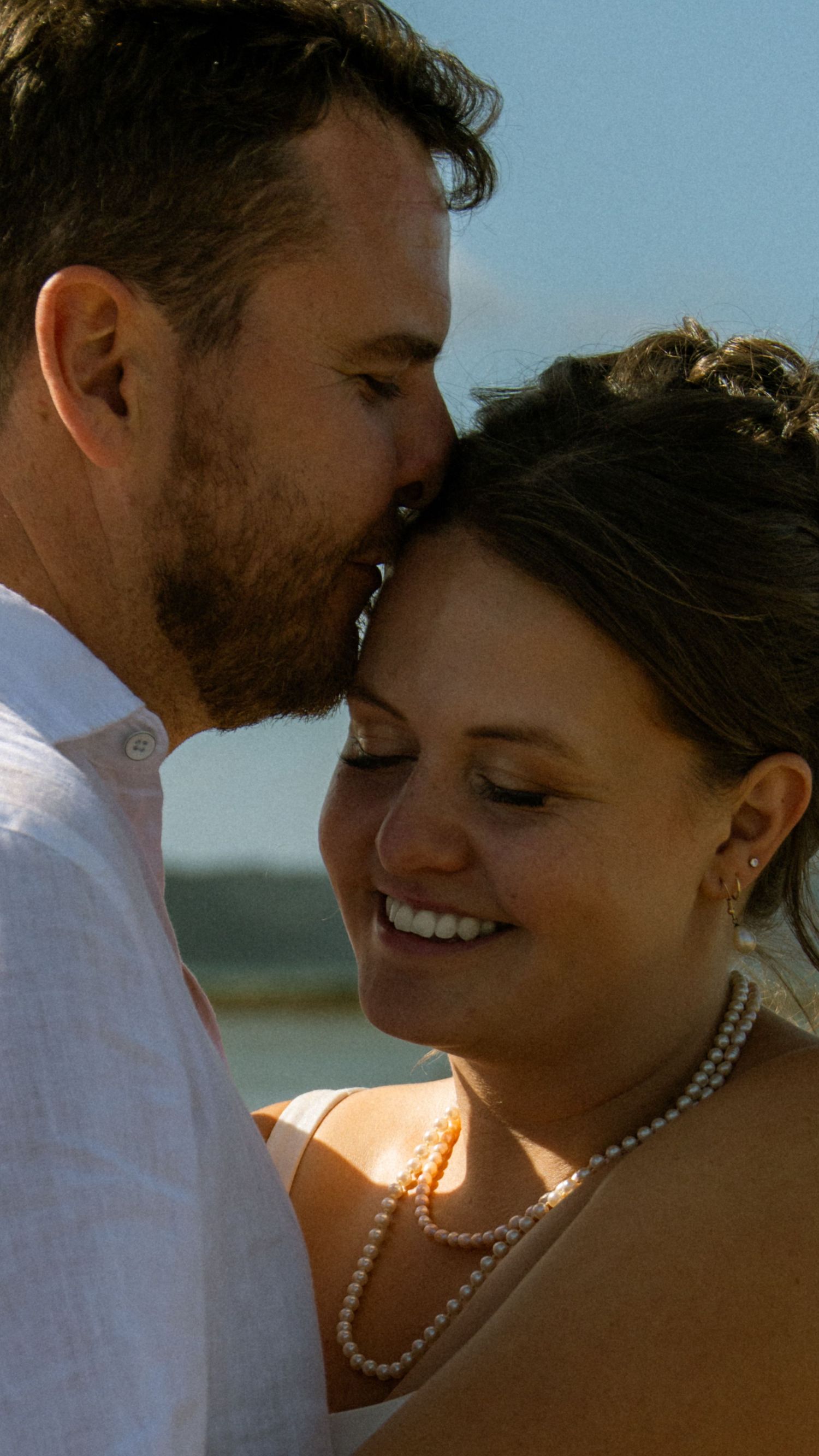 A groom kisses his smiling bride on the forehead at a beach wedding, she wears a pearl necklace.