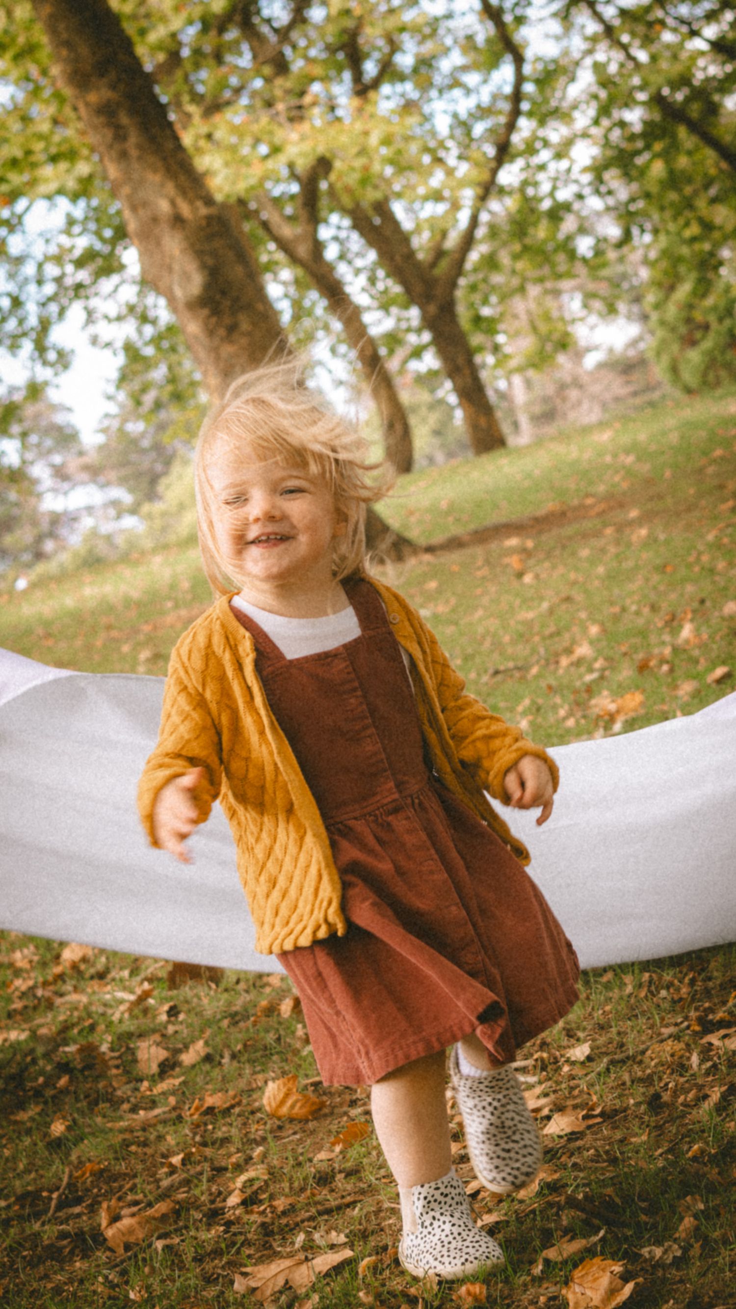 Joyful toddler girl in mustard cardigan and rust dress playing outdoors among autumn leaves in a park.