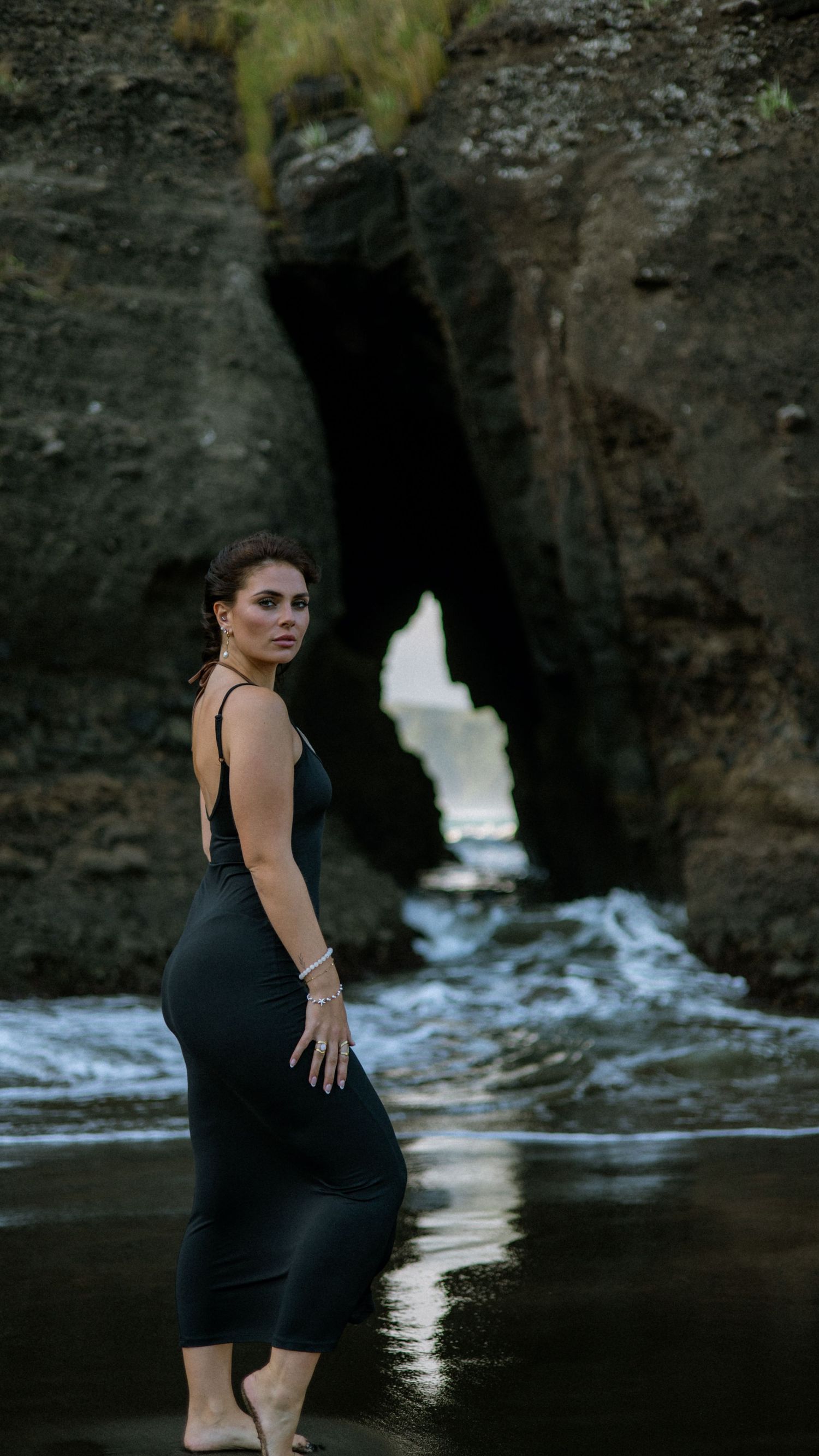 Elegant figure in black dress poses near rocky coastline with ocean waves in background.