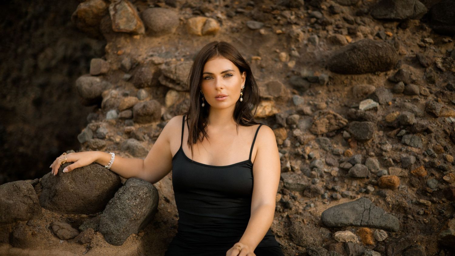 A woman in a black dress poses against a rocky cliff wall in an artistic outdoor photoshoot.