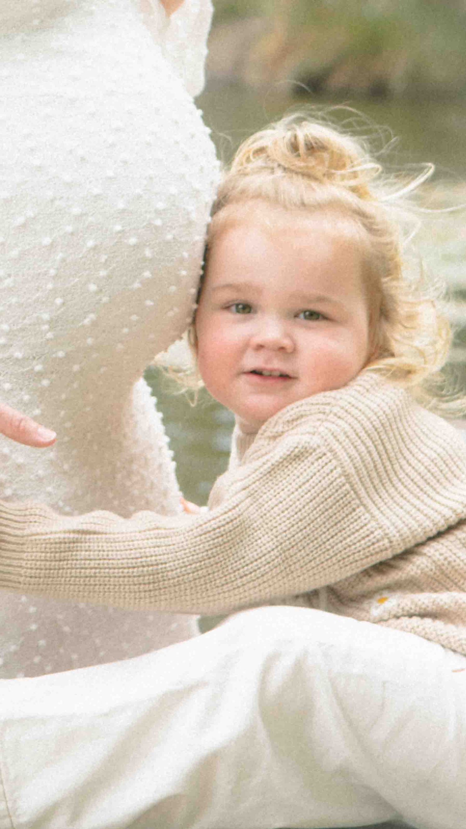 Smiling toddler with wispy blonde hair in a beige knit sweater leans against a pregnant woman's belly in a white dress by a calm stream.