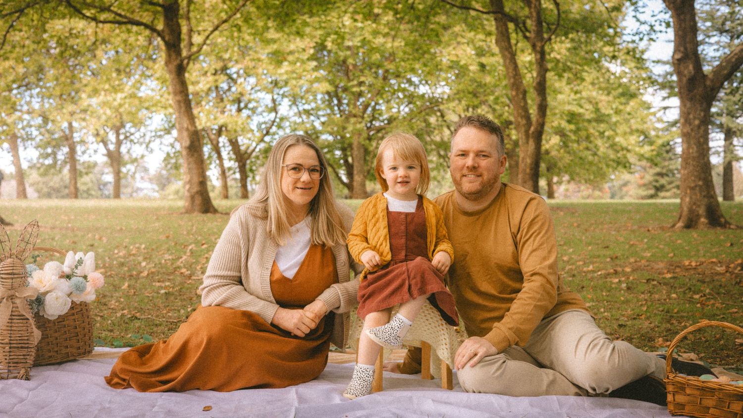 Family of three sitting on a blanket in an autumn park, parents smiling with young toddler in orange dress.