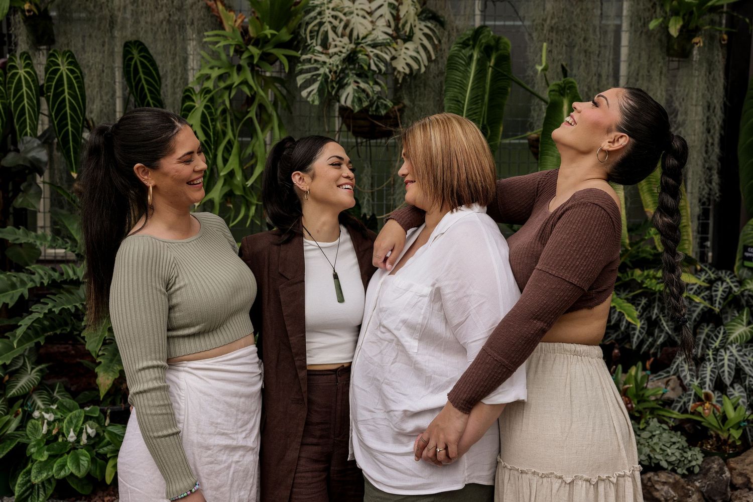 A family in neutral-toned dresses poses together in front of a greenhouse with lush plants.