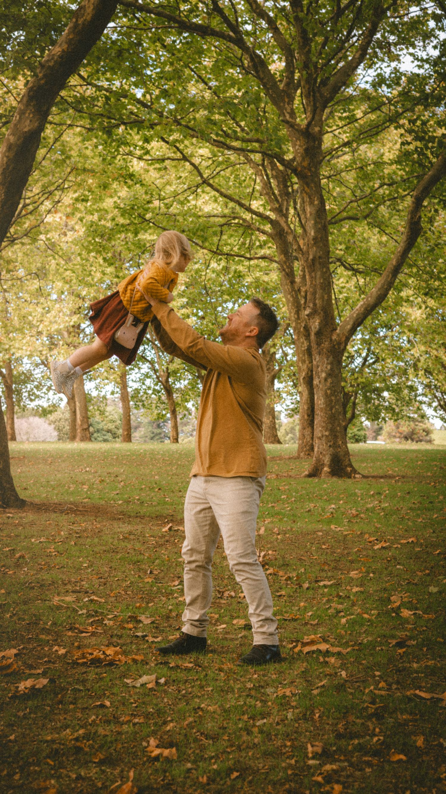 A father joyfully lifts his young child in the air at a lush green park during autumn.