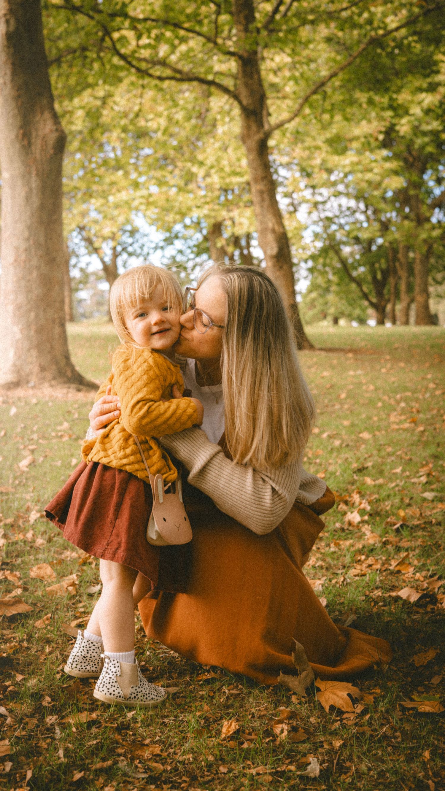 A mother kisses her toddler daughter in an autumn park, both wearing warm fall colors among fallen leaves.