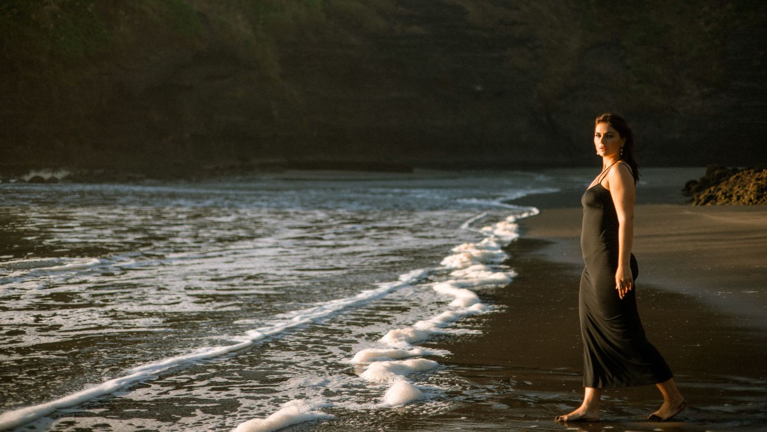 A figure in a white dress dances gracefully on a beach at sunset with mountains in the background.
