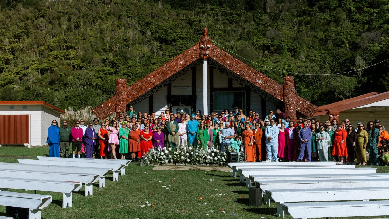 A panoramic view of a rustic outdoor wedding ceremony setup with white benches and a wooden arch in a garden setting.