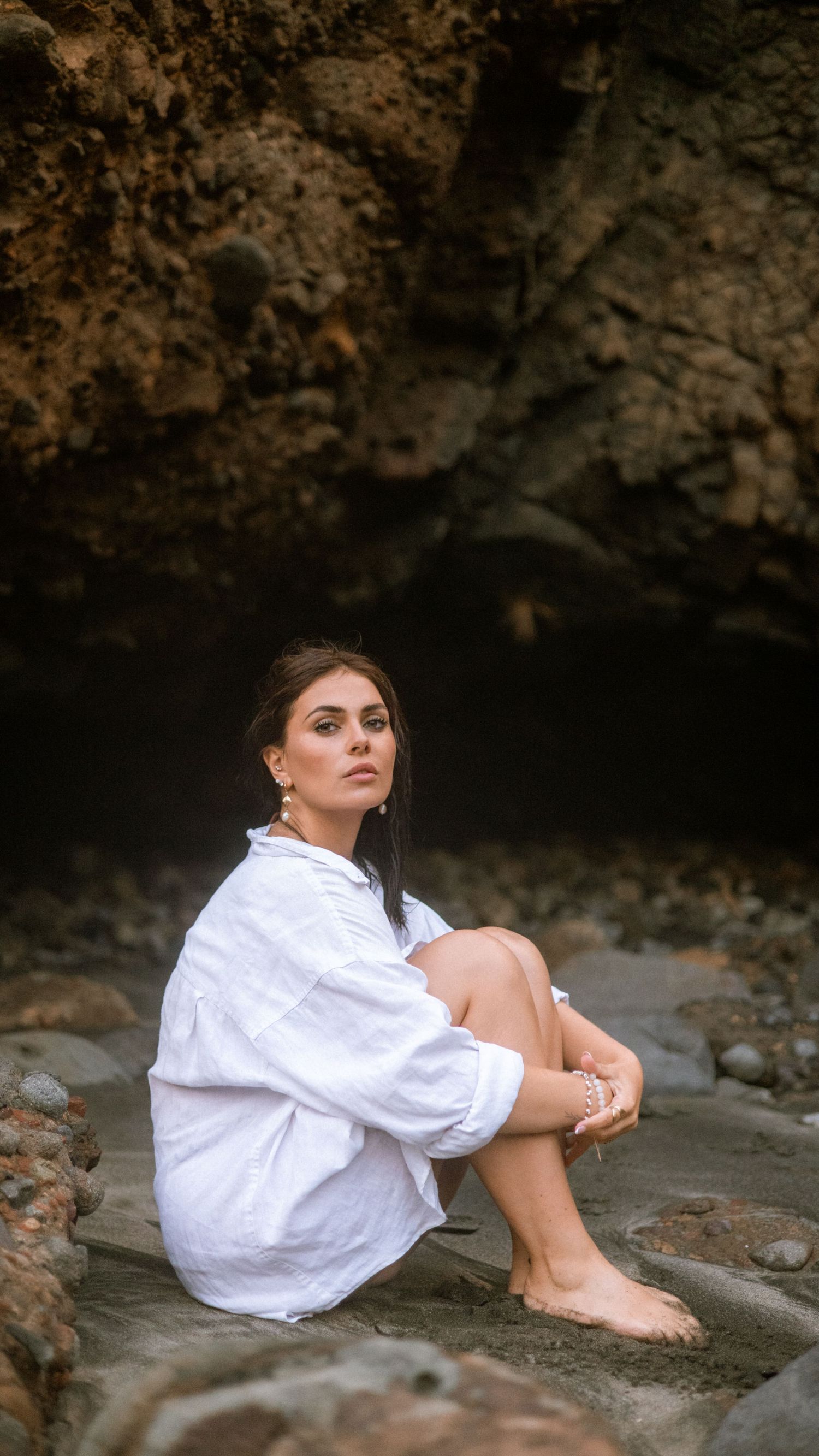 A person in a white shirt sits contemplatively on rocky terrain against a dark cave-like background.