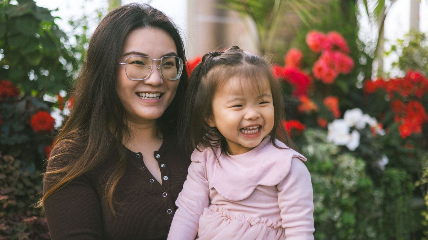 A smiling Asian woman holding a laughing toddler girl in a pink jacket, posing outdoors with colorful flowers.