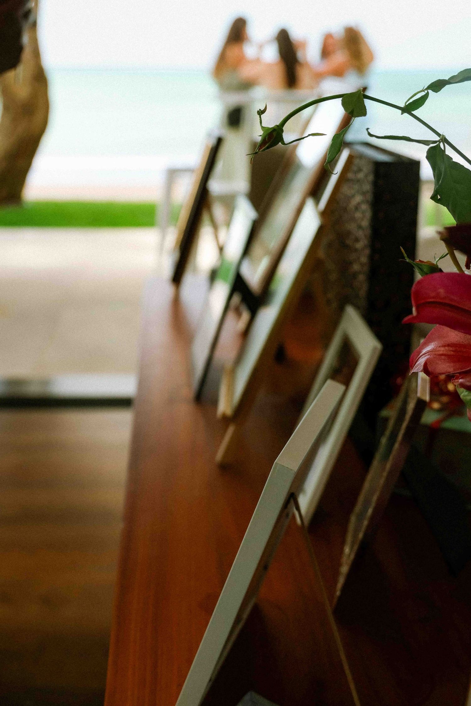 Framed photos displayed on a wooden table with tropical flowers, overlooking a beachside gathering of women in the background.