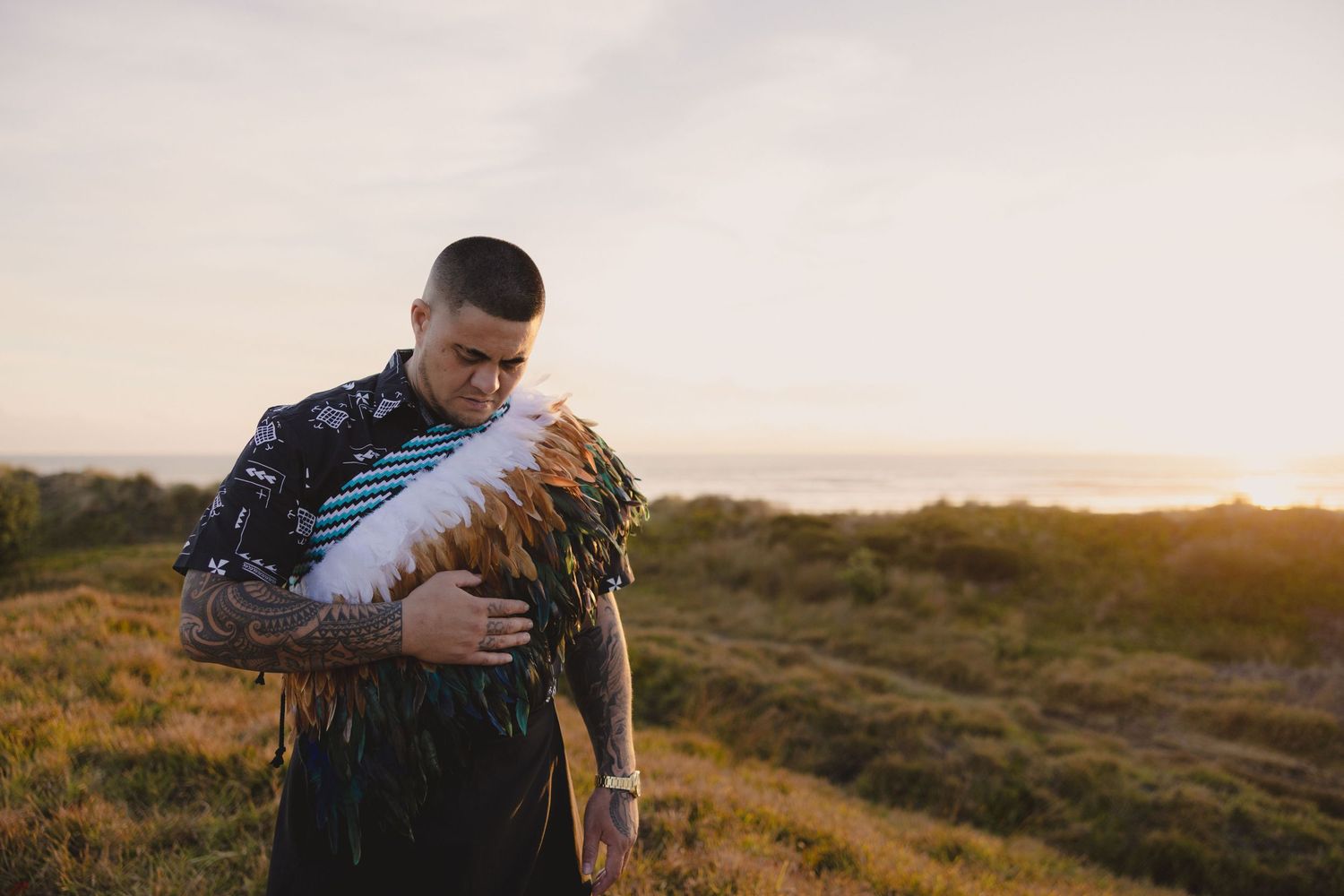 Someone in a dark sweater holds a chicken while standing on a grassy hill at sunset by the ocean.