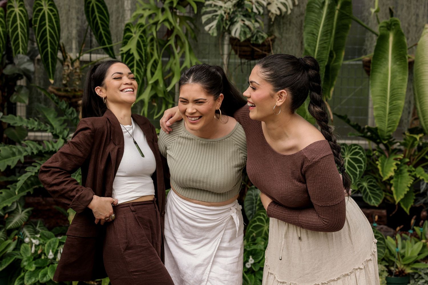 Three sisters share joyful laughter together in an outdoor garden setting wearing earth-toned clothing.