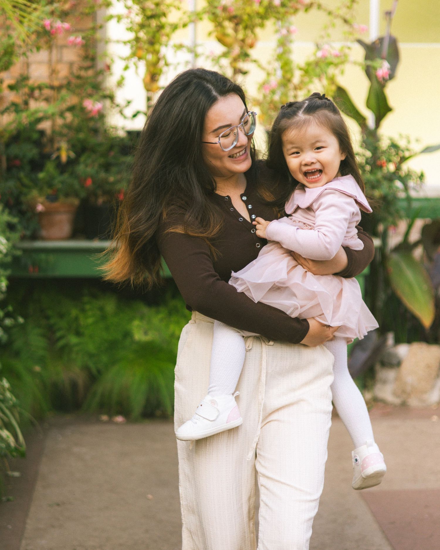A smiling Asian woman in white pants holds a laughing toddler girl in a pink dress outdoors among green plants.