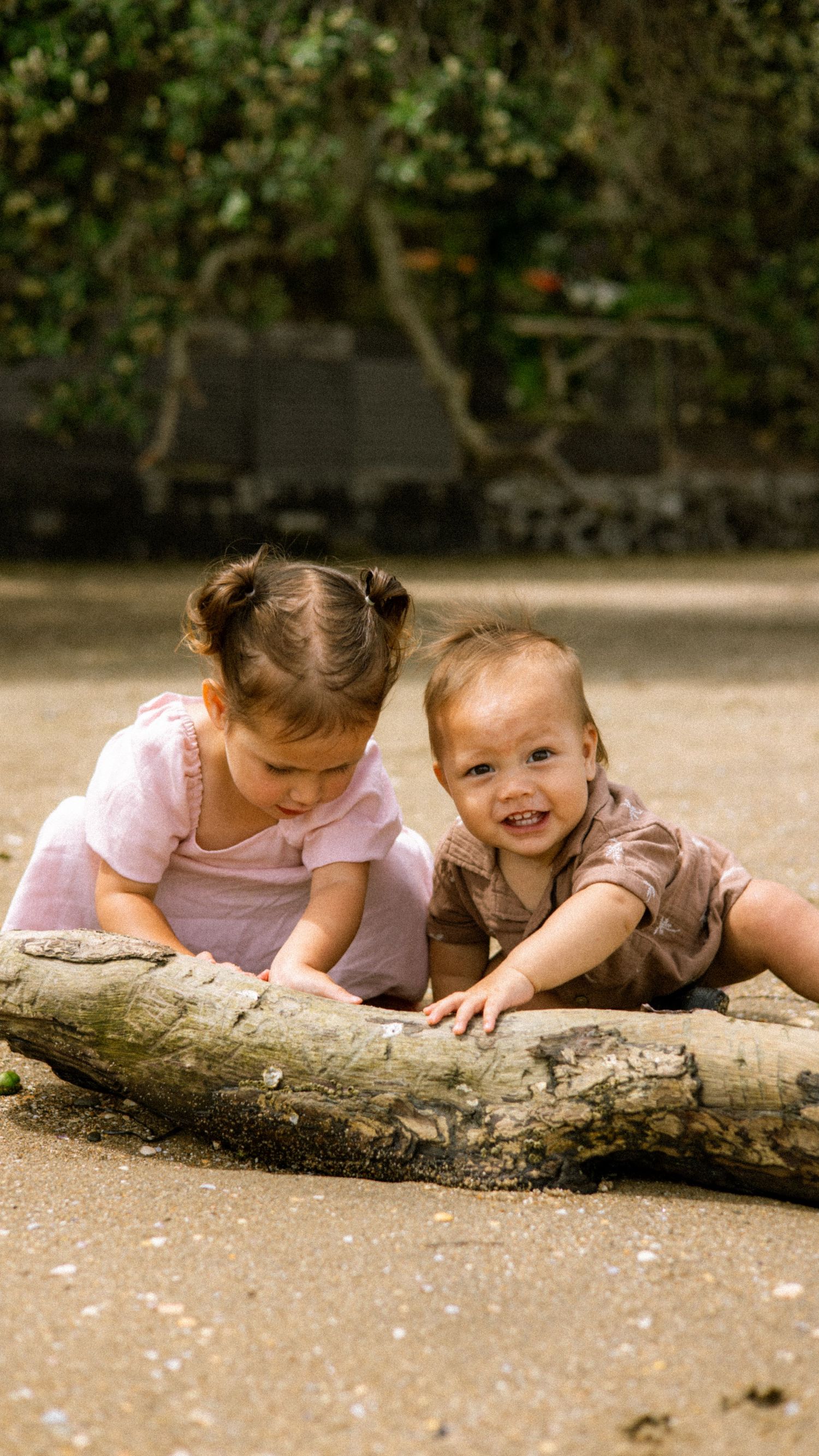 Two young children in pink playing and laughing while lying on a fallen tree trunk outdoors.