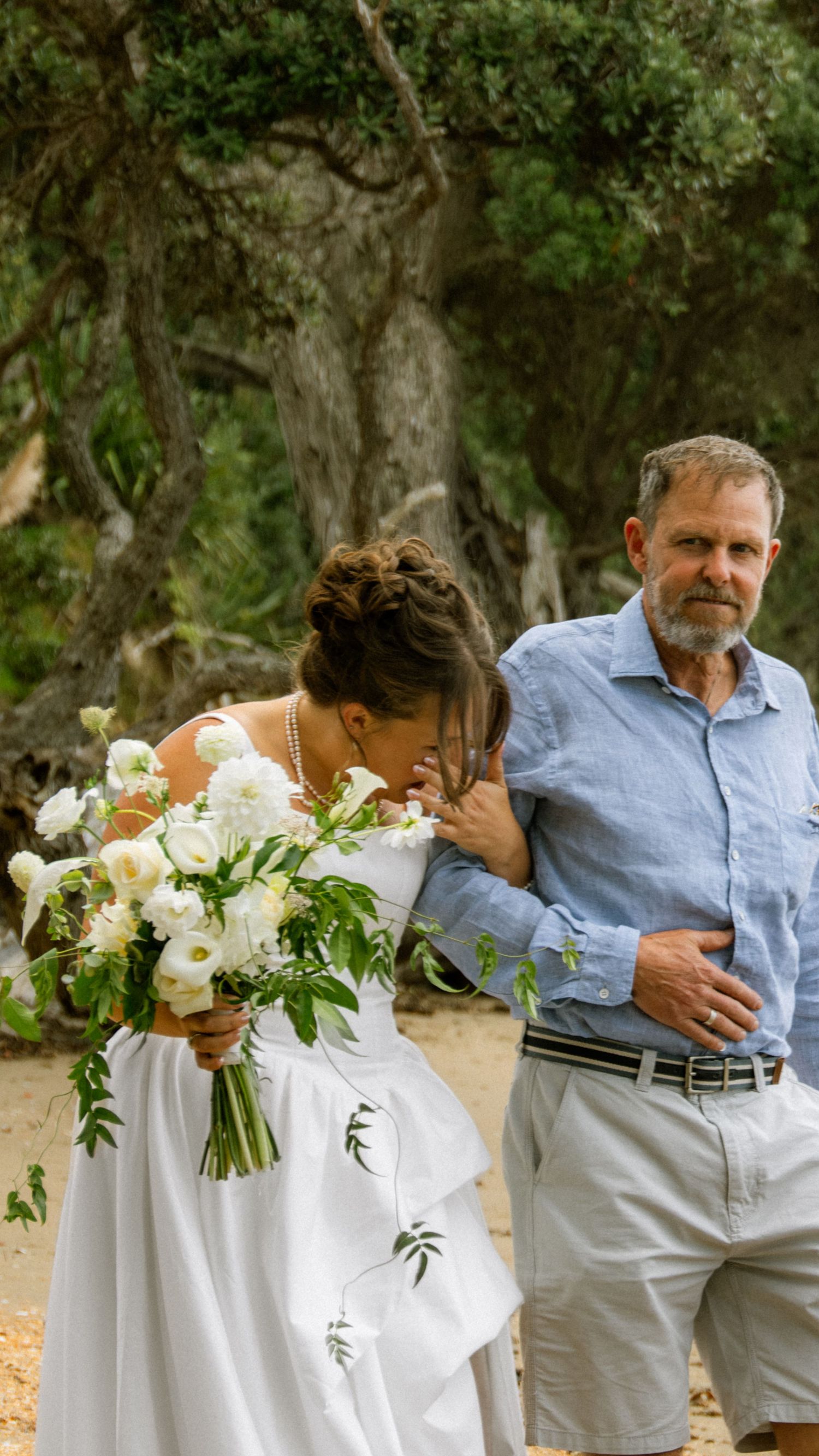 A bride in a white gown holds a lush white bouquet while walking with an older man in a blue shirt outdoors.