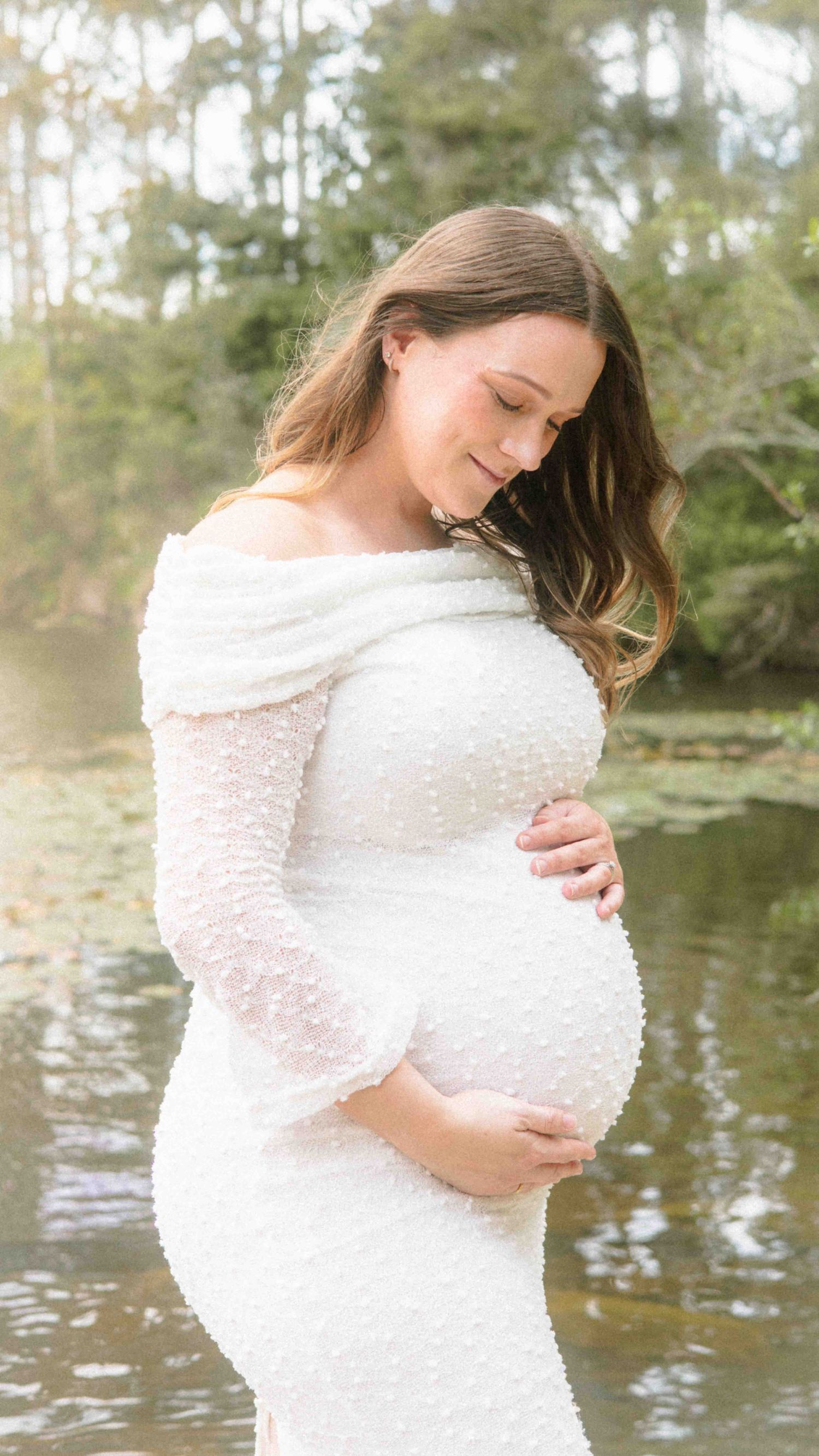 Pregnant woman in a white off-shoulder textured dress cradles her baby bump while standing beside a calm lake in a maternity photoshoot.