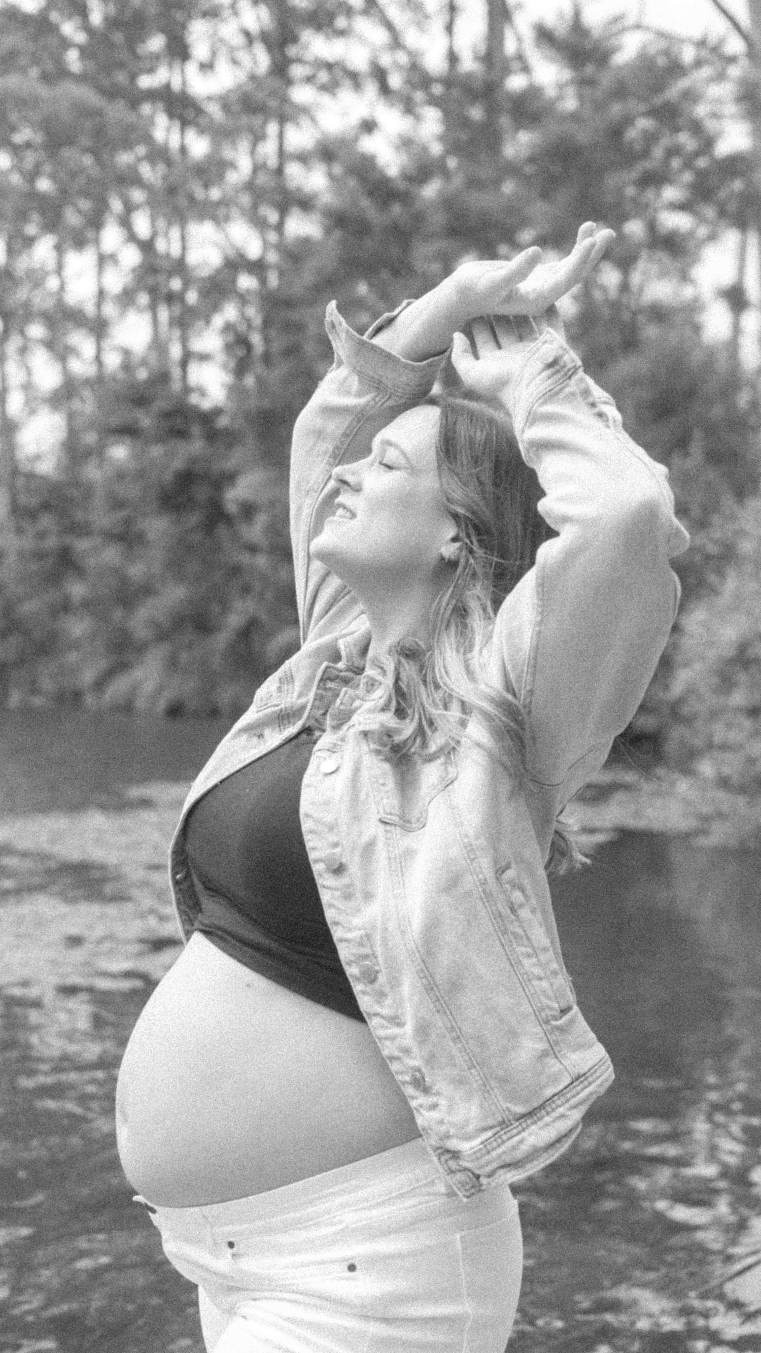 Black and white maternity portrait of a pregnant woman in a denim jacket with arms overhead, eyes closed, standing by a tranquil lake.