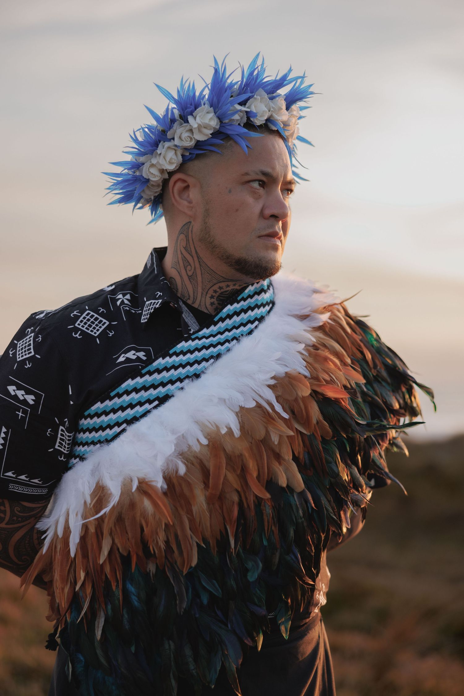 A person wearing a blue flower crown and feathered cloak poses against a sunset backdrop.