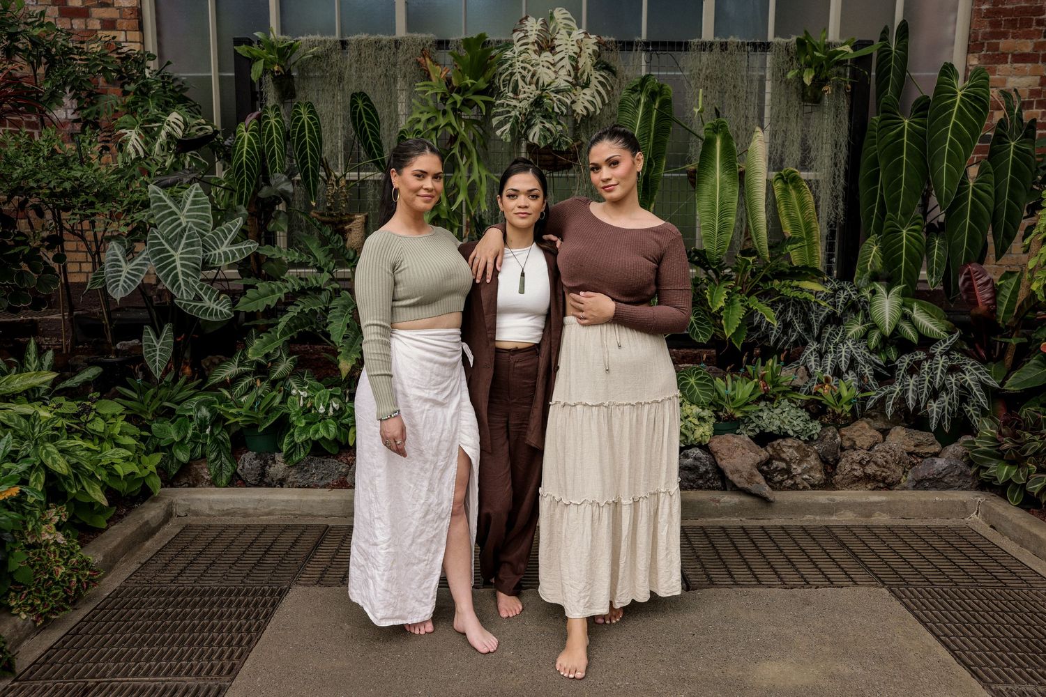 Three people in neutral tones posing in front of greenhouse windows.