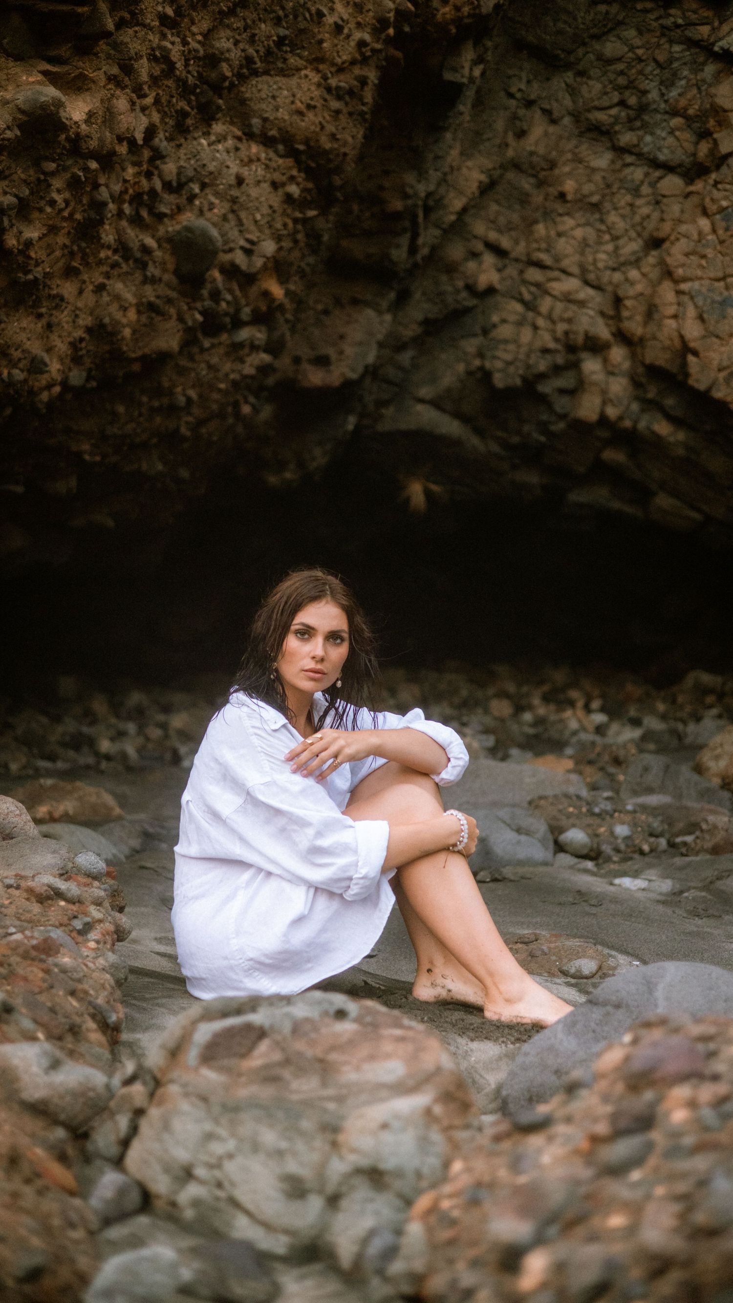 Person in white dress sits thoughtfully among rocky coastal cave formations at sunset.