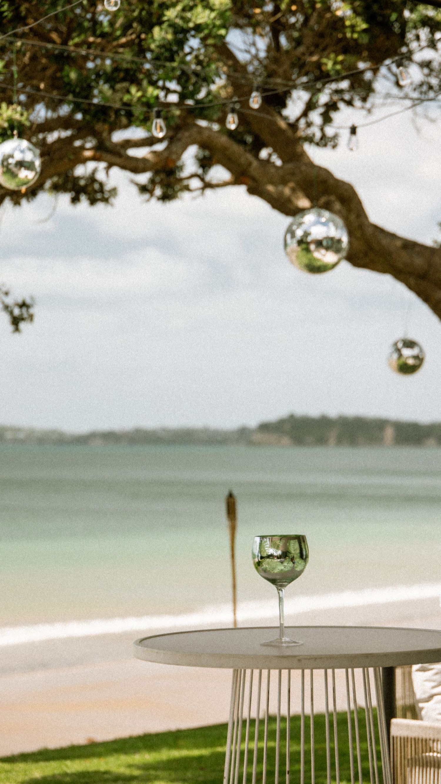 A wine glass on a beachside table under a tree with disco balls, overlooking calm turquoise waters.