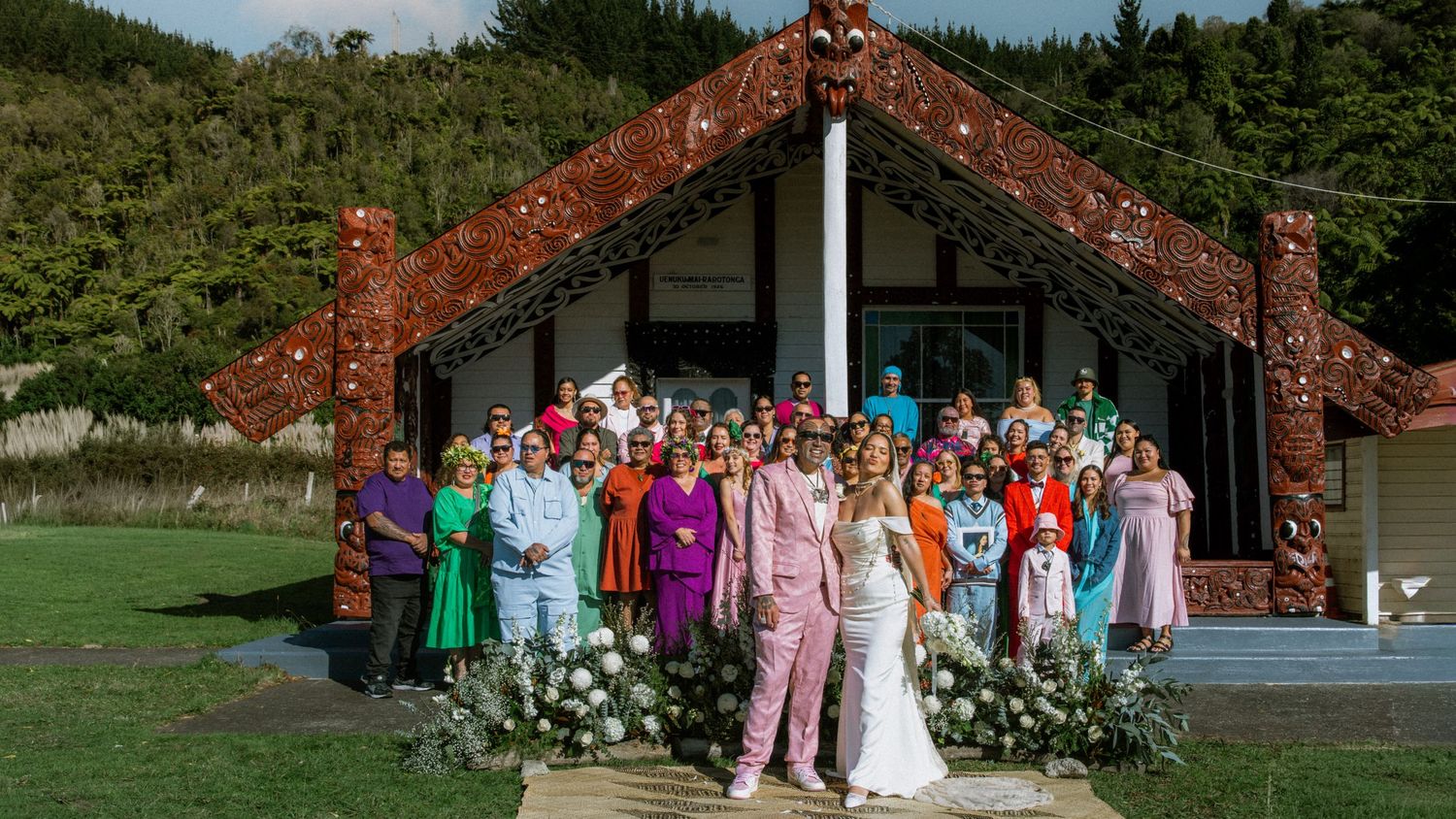 Wedding party gathered in front of traditional Maori meeting house with ornate wooden carvings in New Zealand.