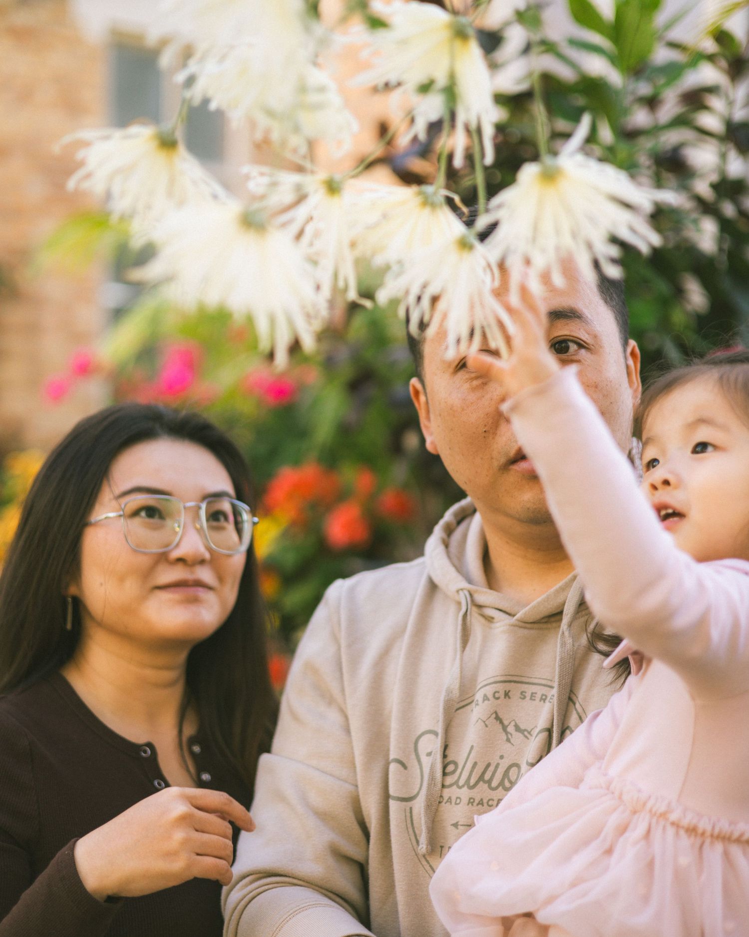 A family of three admires white flowers outdoors, with a young child reaching up to touch the blooms.
