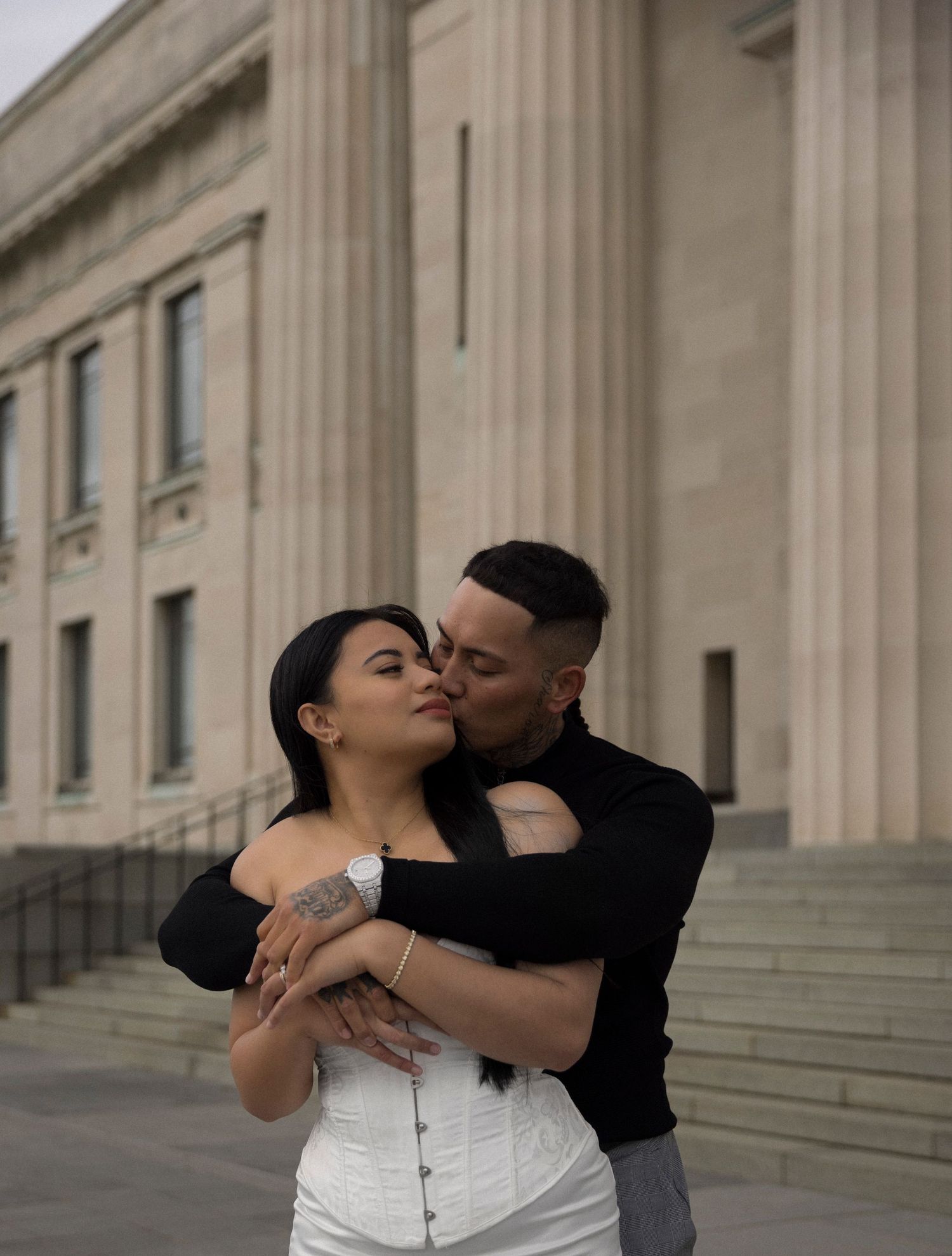 A romantic couple shares tender kisses and embraces on the grand stone steps of a historic building with columns.