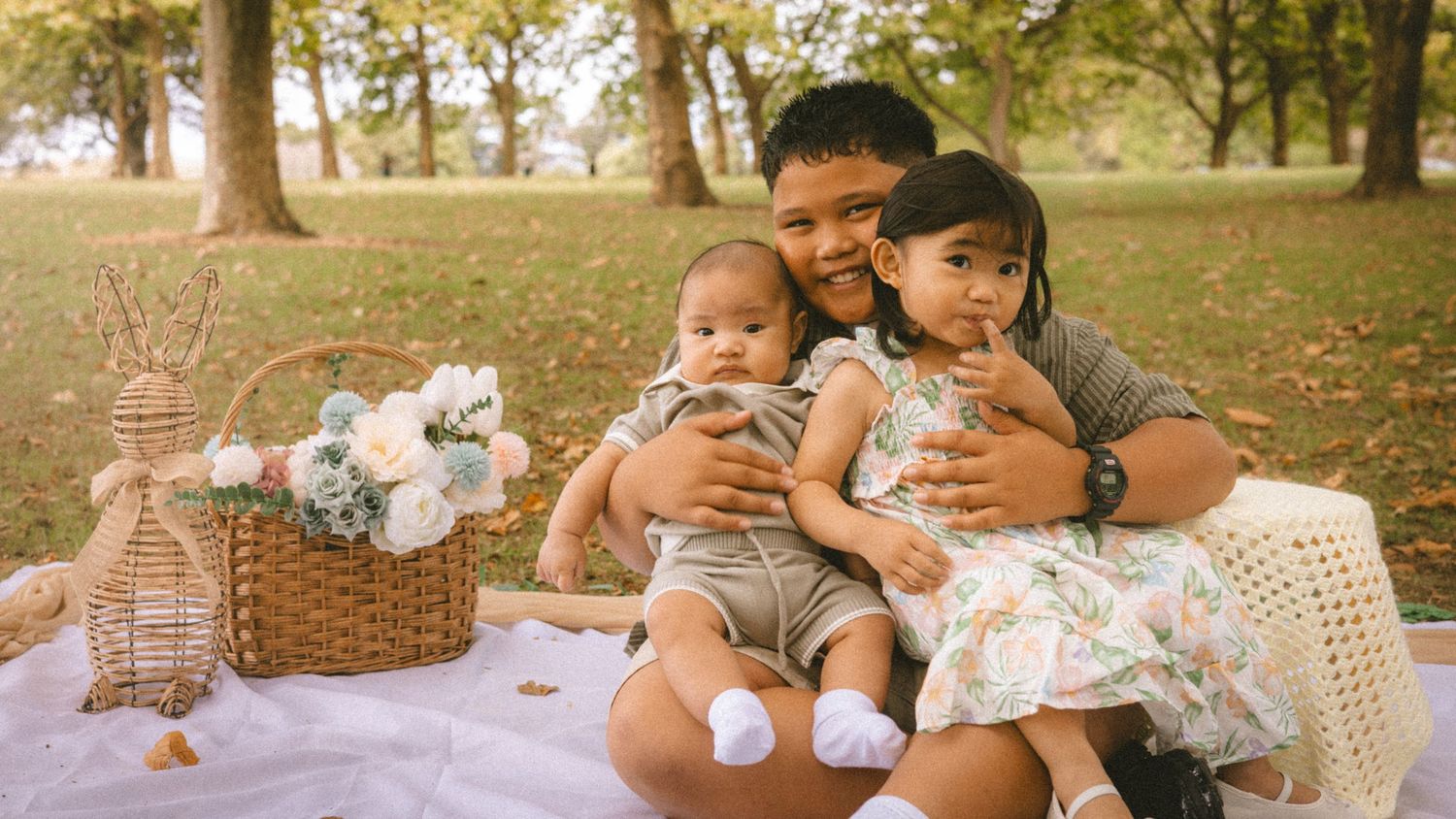 A young boy sits on a picnic blanket in a park, holding two babies, with a wicker basket and flowers nearby.
