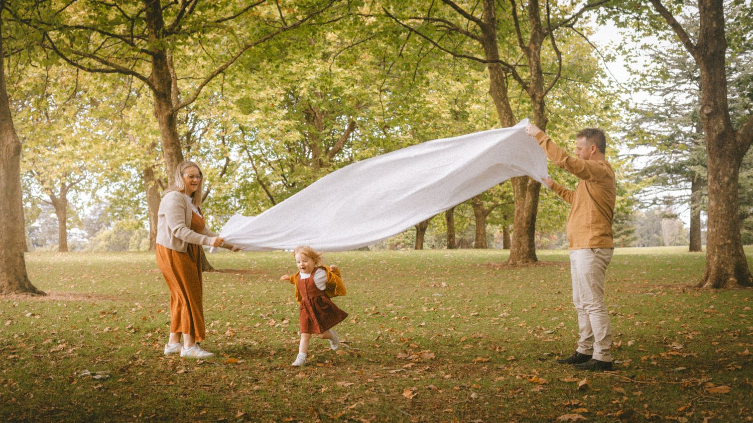 Family spreading white blanket in autumn park with trees and green grass on a sunny day.