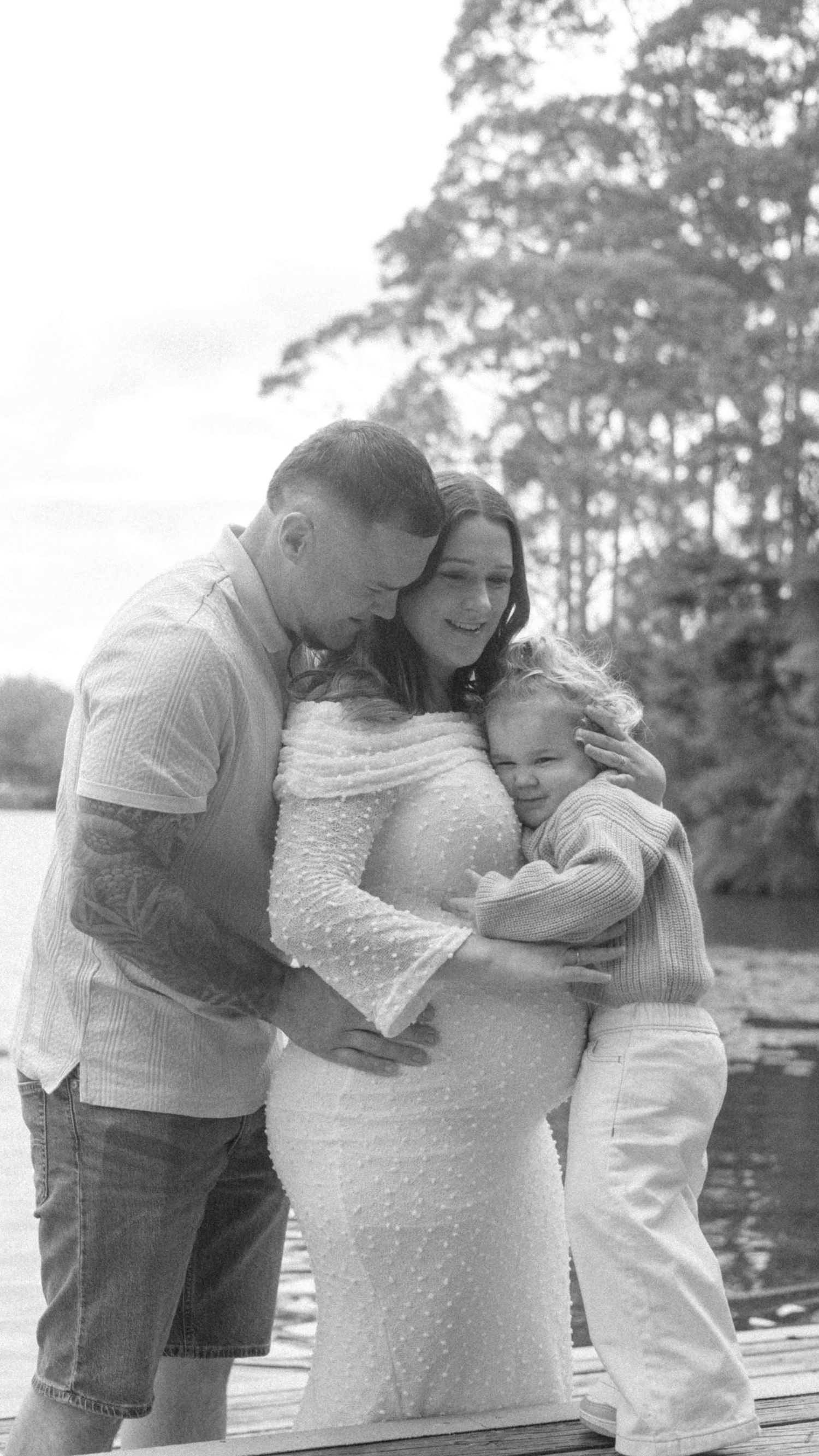 Black and white maternity photo of an expectant couple embracing their young child on a wooden dock beside a lake with tall trees.